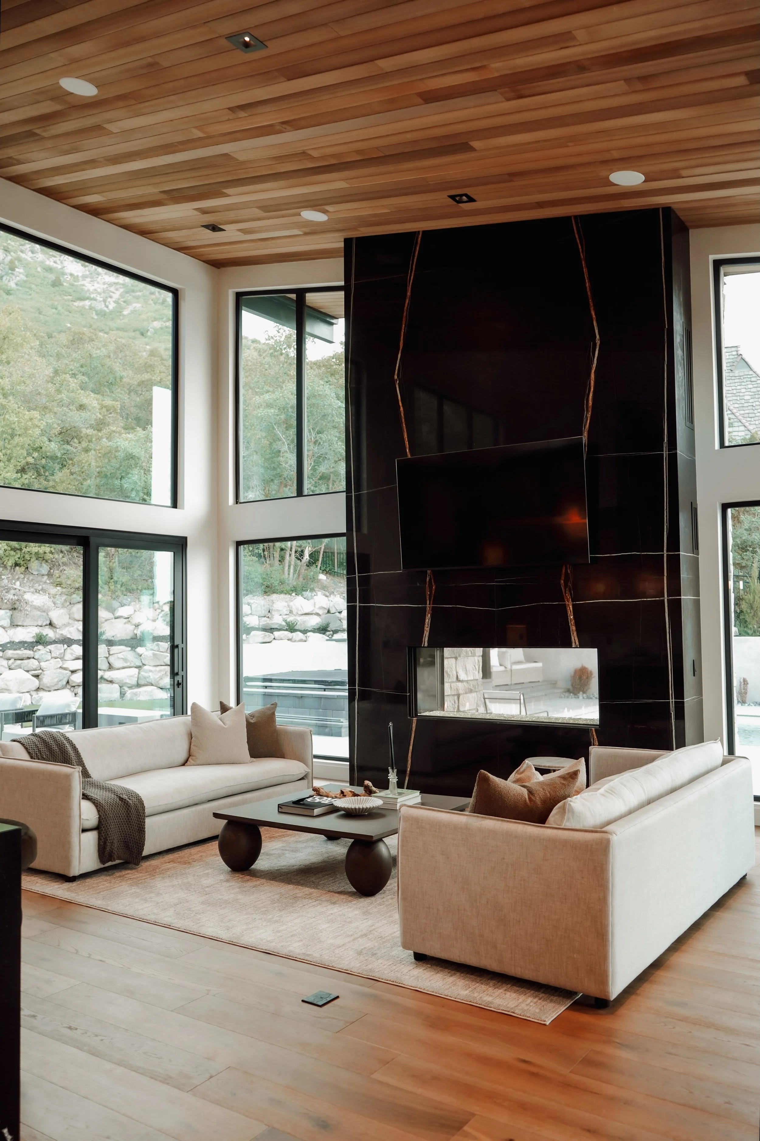Living room with large floor-to-ceiling windows, two beige sofas with pillows, a black coffee table, and a modern black marble fireplace with a mounted TV. Wooden ceiling and floor, outside view of trees and rocks.