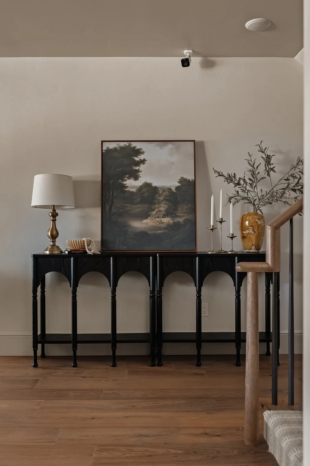 A black console table against a white wall with a landscape painting, a white table lamp, a decorative bowl, a beige and brown vase with foliage, and silver candlesticks with white candles on top.