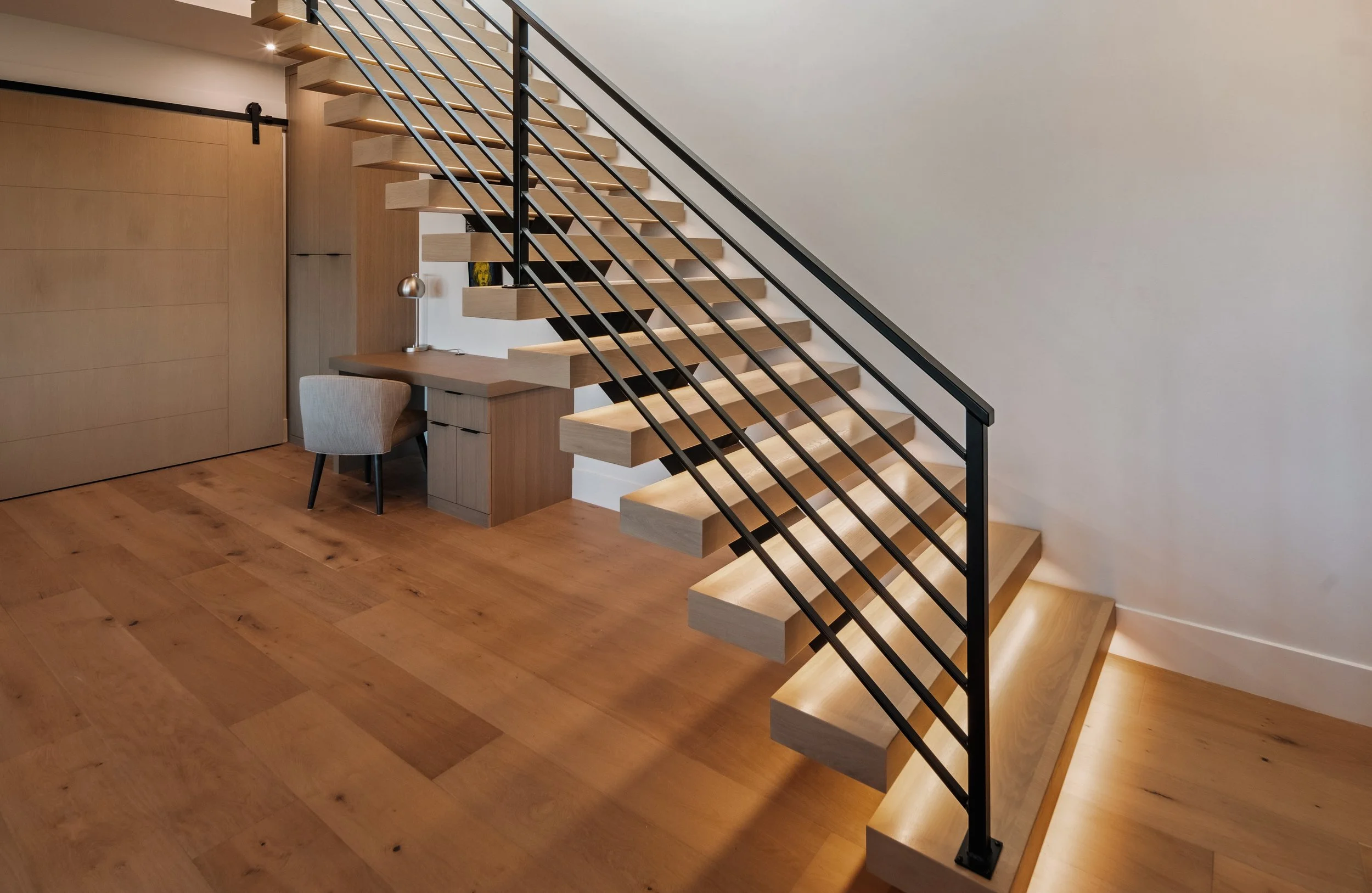 Interior of a modern home featuring a floating wooden staircase with black metal railings, a wooden floor, and a desk with a chair underneath the stairs.