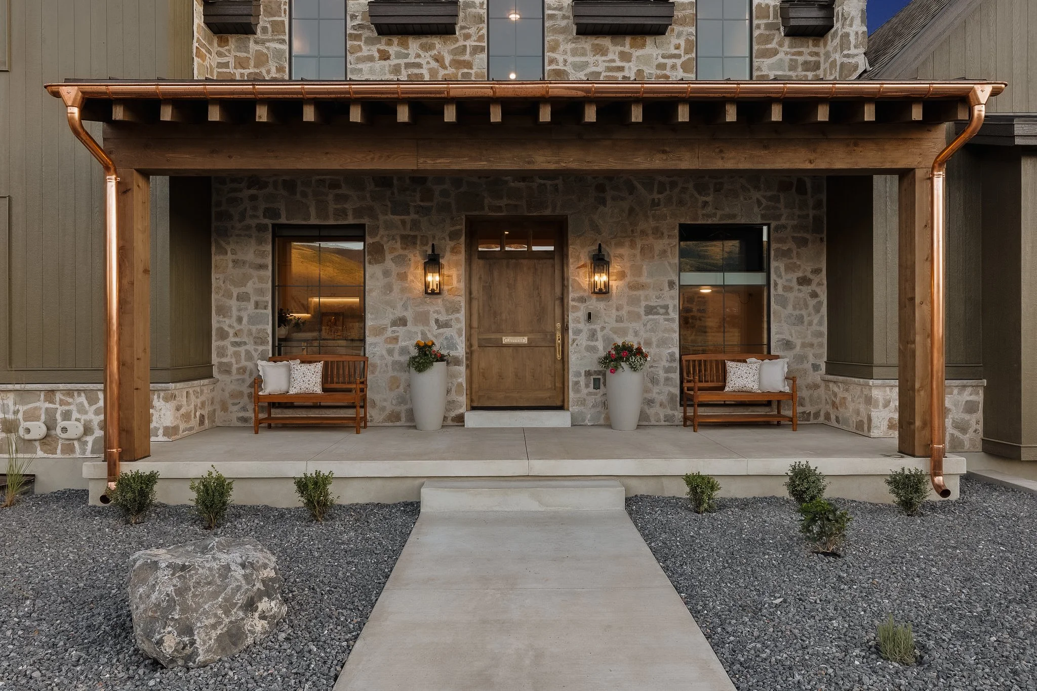 Front porch of a house with a stone and wood facade, two wooden benches with cushions, large white planters with flowers, and two wall-mounted lanterns beside the main door. Gravel and a concrete walkway lead to the entrance.