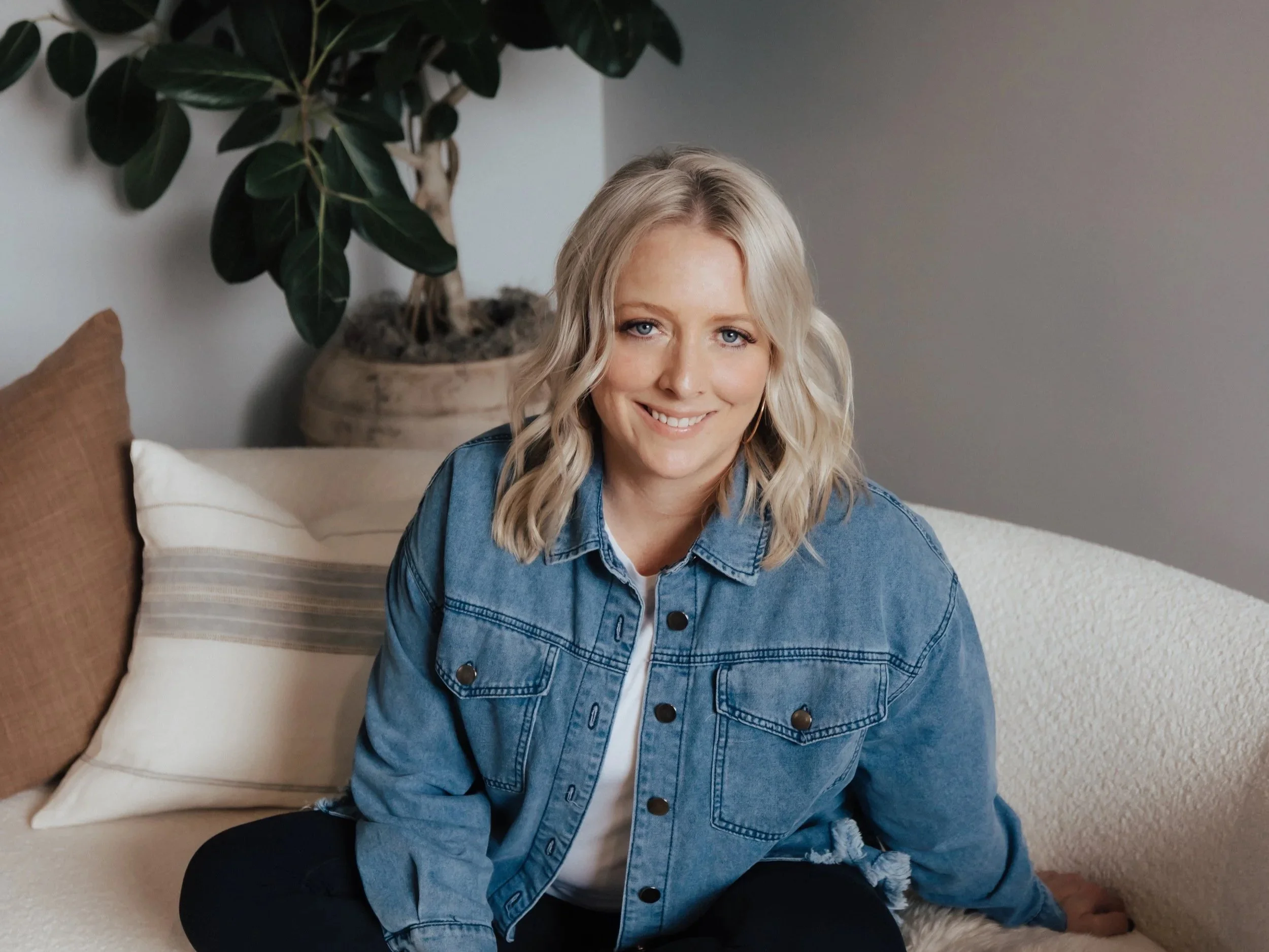 A smiling woman with blonde hair sitting on a beige couch, wearing a denim jacket and black pants, with a large potted plant and decorative pillows in the background.