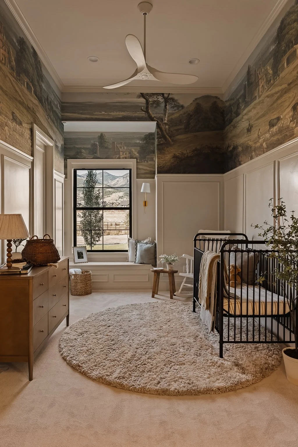 A cozy nursery with beige carpet, round shaggy rug, black metal crib, wooden dresser with lamp, wall mural of countryside, window seat with cushions, small side tables with plants and framed photo, ceiling fan, and large window showing a landscape wi