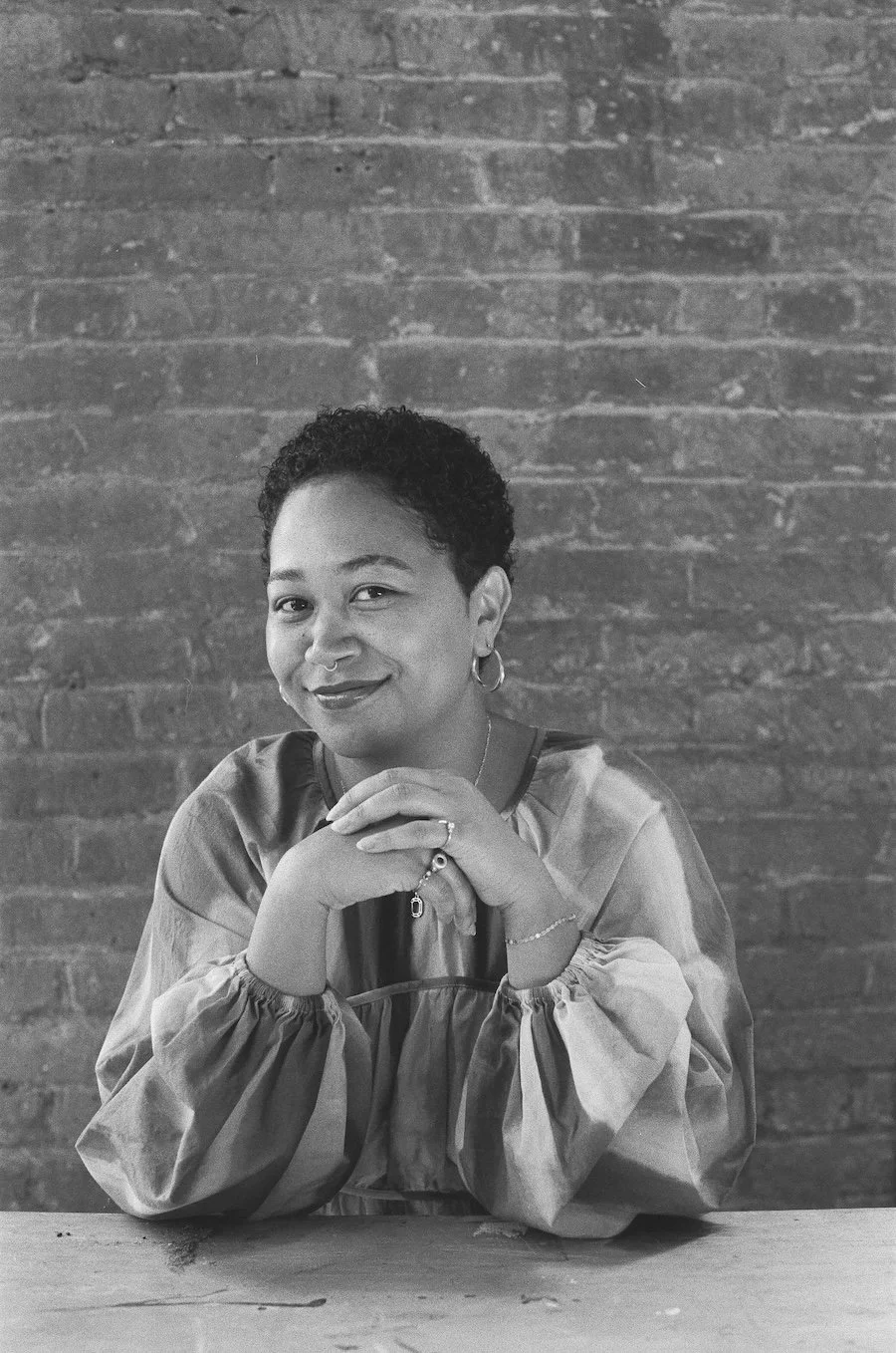 Black and white photo of a Anela Malik sitting at a table with her hands clasped, smiling, wearing a long-sleeve top and earrings, with a brick wall in the background.