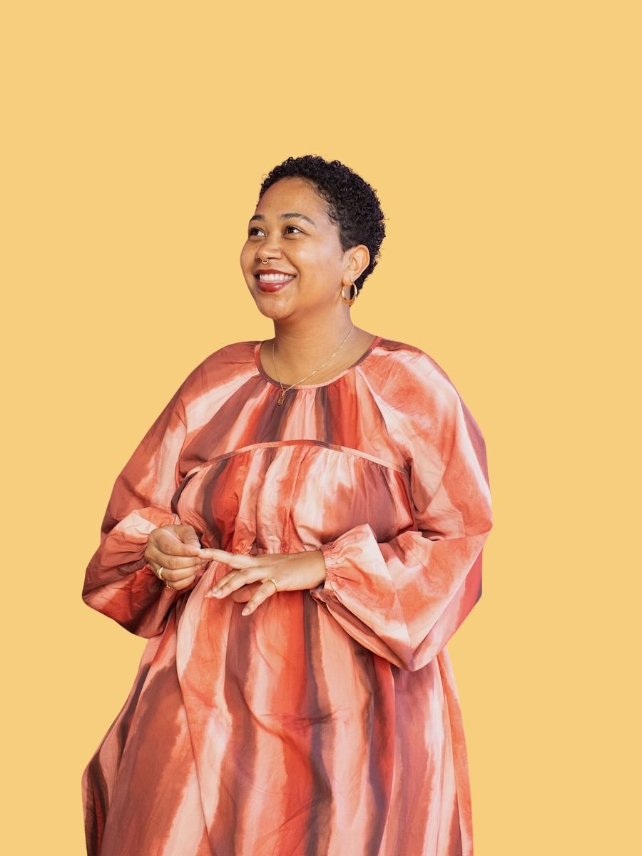 Anela Malik with short curly hair and earrings is smiling, wearing a multicolored dress with shades of orange, red, and pink, standing against a yellow background.