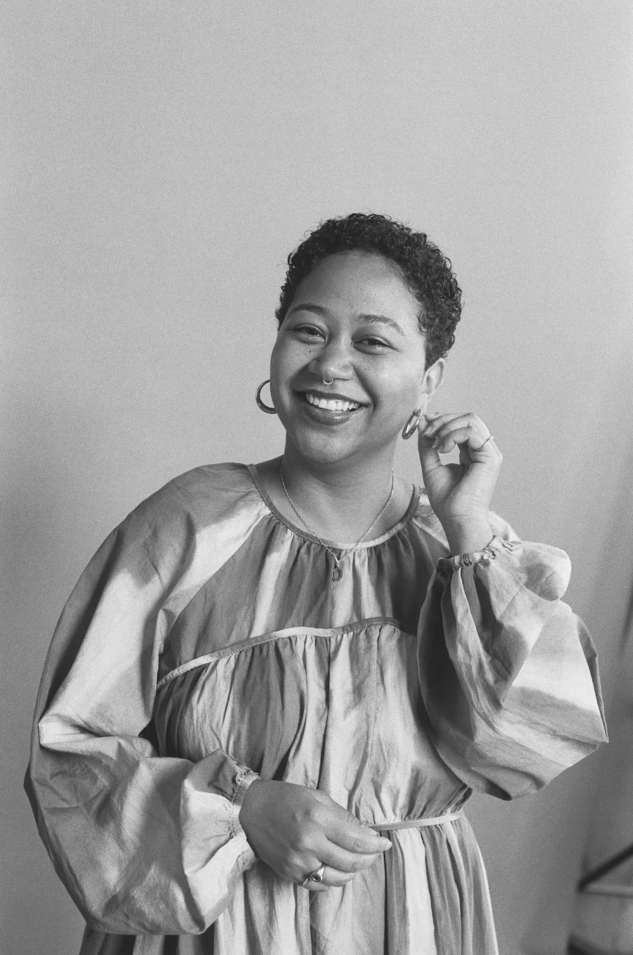 A black and white photograph of Anela Malik with short curly hair smiling, wearing a loose, light-colored dress with puffy sleeves, earrings, a nose ring, and rings, standing against a plain background.