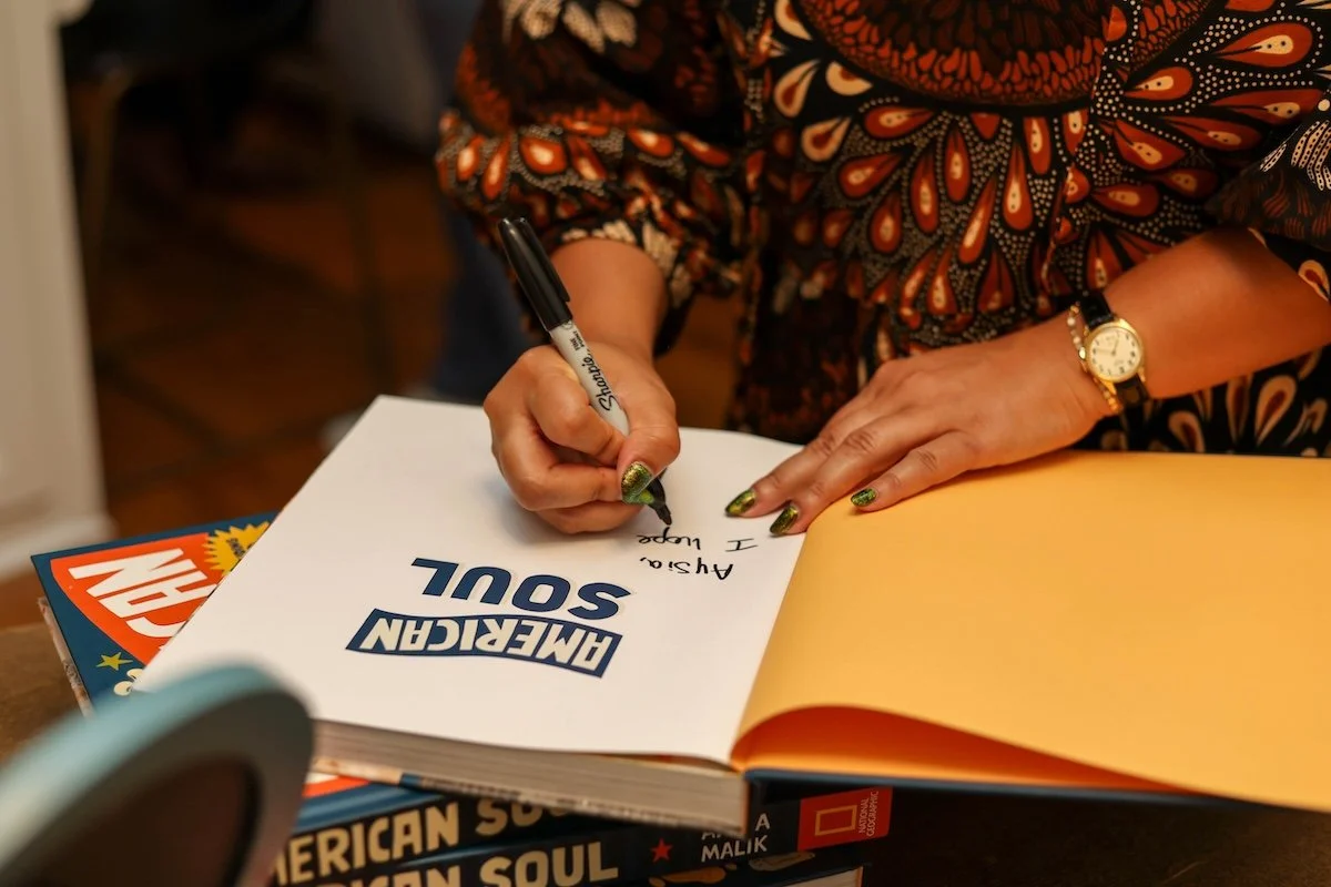 Anela Malik signing a book titled 'American Soul' with a black Sharpie marker. She is wearing a patterned blouse, a gold watch, and has decorated nails. The book is open on a table, with other colorful books stacked underneath.