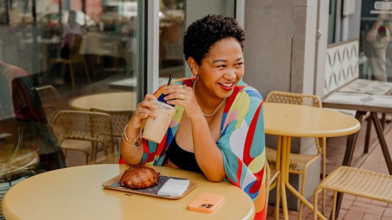 Anela Malik with short curly hair enjoying a drink and pastry at a cafe, sitting at a yellow table outside.