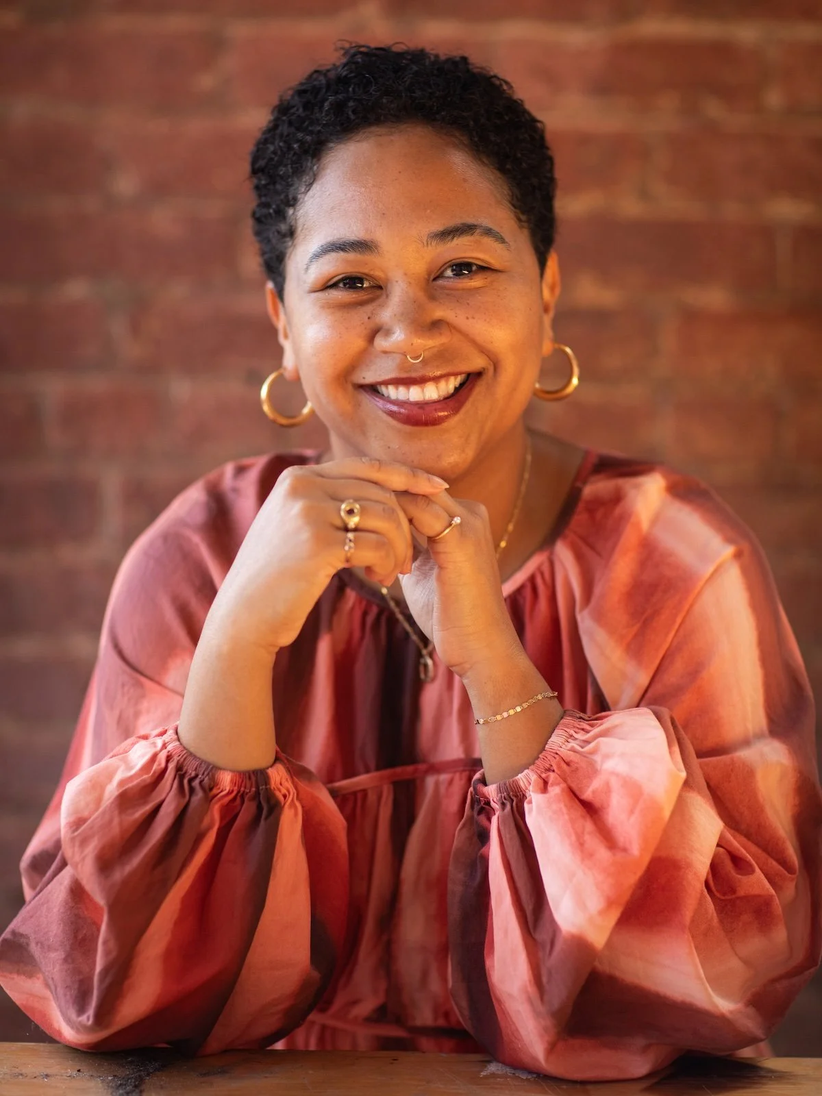Anela Malik with short curly hair and a septum piercing smiling at the camera, wearing gold hoop earrings, rings, and bracelets, sitting at a table with a brick wall background.