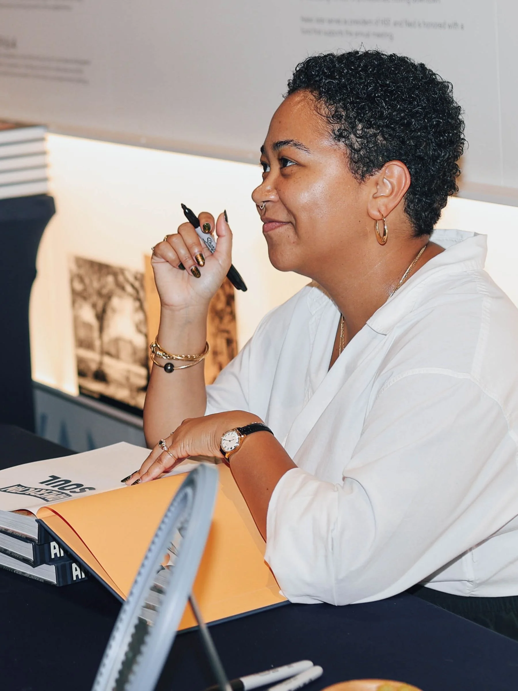 Anela Malik with short curly hair, wearing a white shirt, gold hoop earrings, a watch, and bracelets, is sitting at a table with copies of her book, holding a black marker, in an indoor setting.
