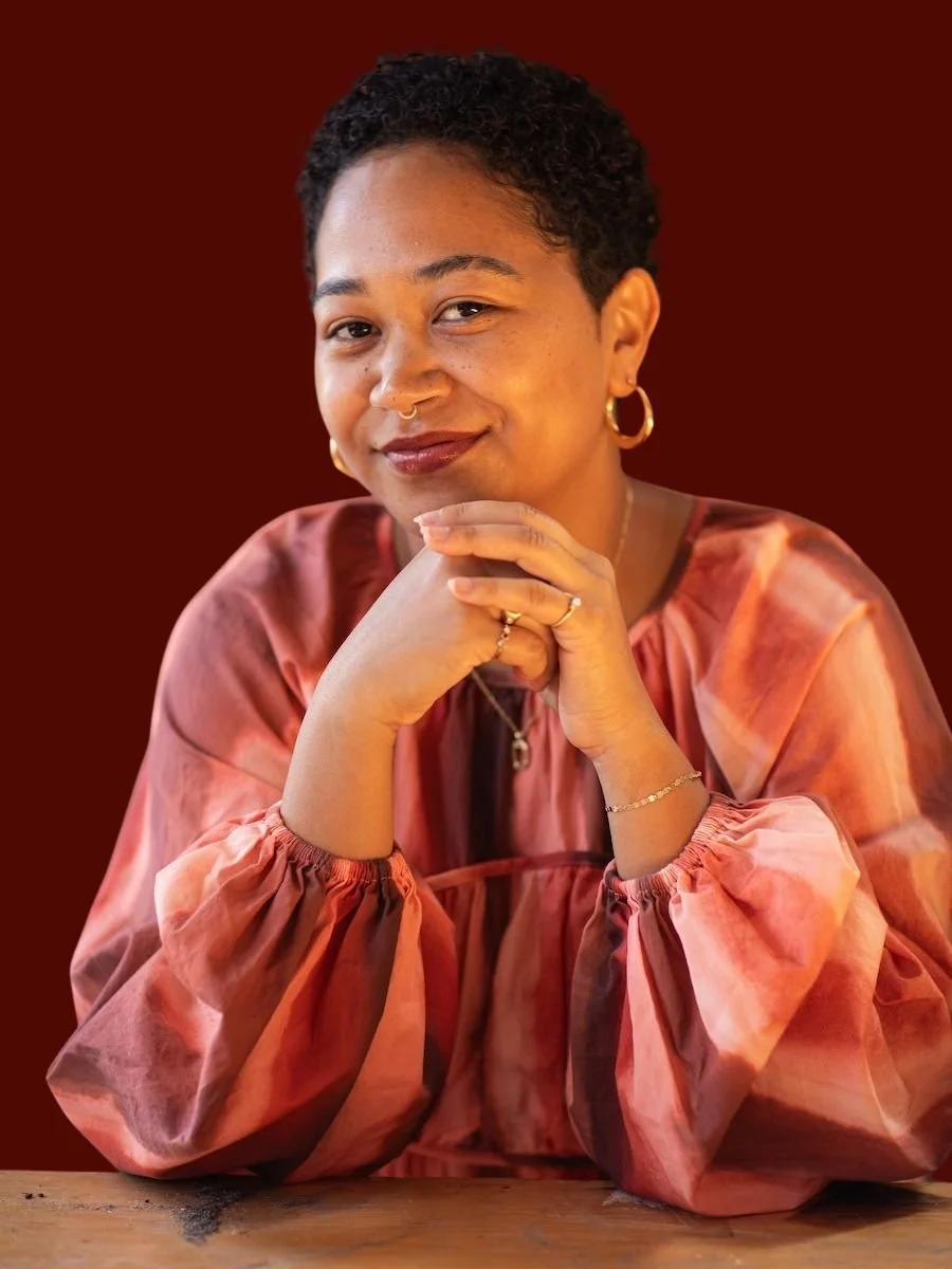 Portrait of a Anela Malik with short curly hair wearing a pink tie-dye blouse, gold hoop earrings, and rings, sitting at a wooden table with a dark red background.