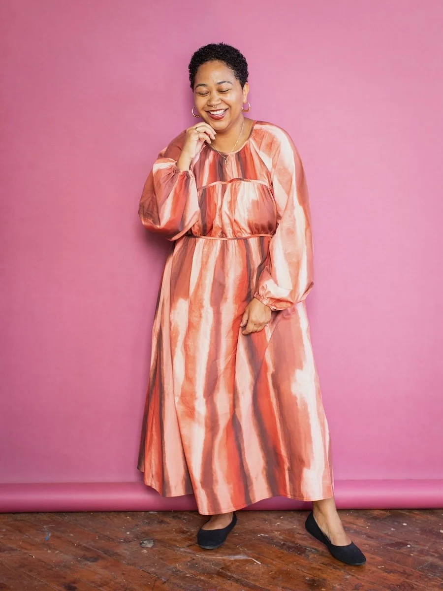 Anela Malik with short curly hair wearing a flowing, multicolored orange and pink striped dress, smiling and standing against a pink background, with one hand near her face and the other resting on her hip.