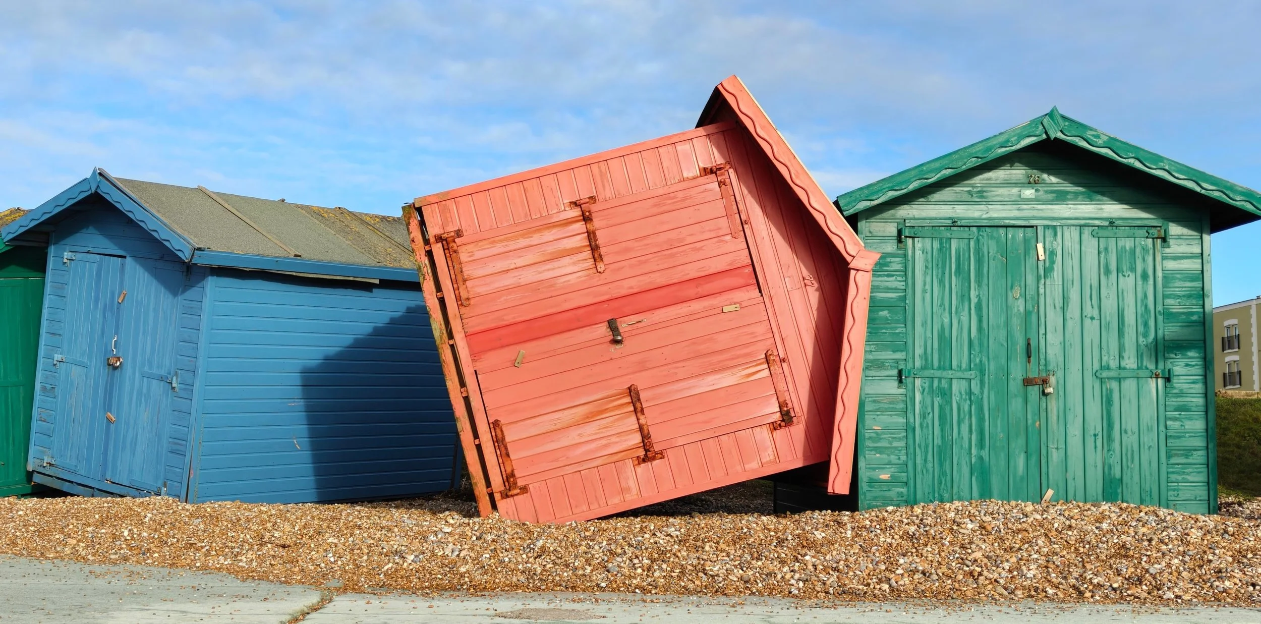 Colorful beach huts, with one hut tipping over, painted in blue, pink, and green, set on a gravel surface under a partly cloudy sky. Free mortgage and insurance advice