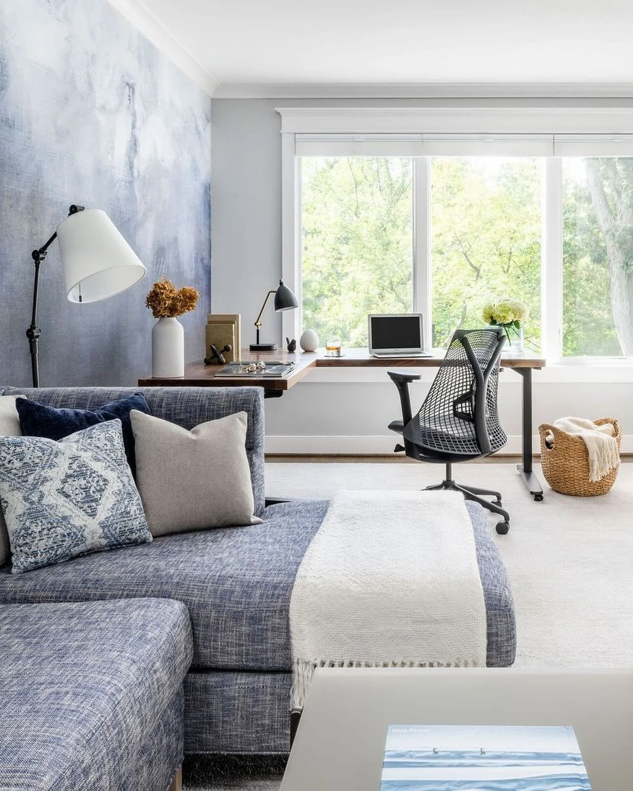 A cozy living room and home office space with a blue sofa adorned with decorative pillows, a white throw blanket, a desk with a computer, desk lamp, and decorative items near a large window showing green trees outside.