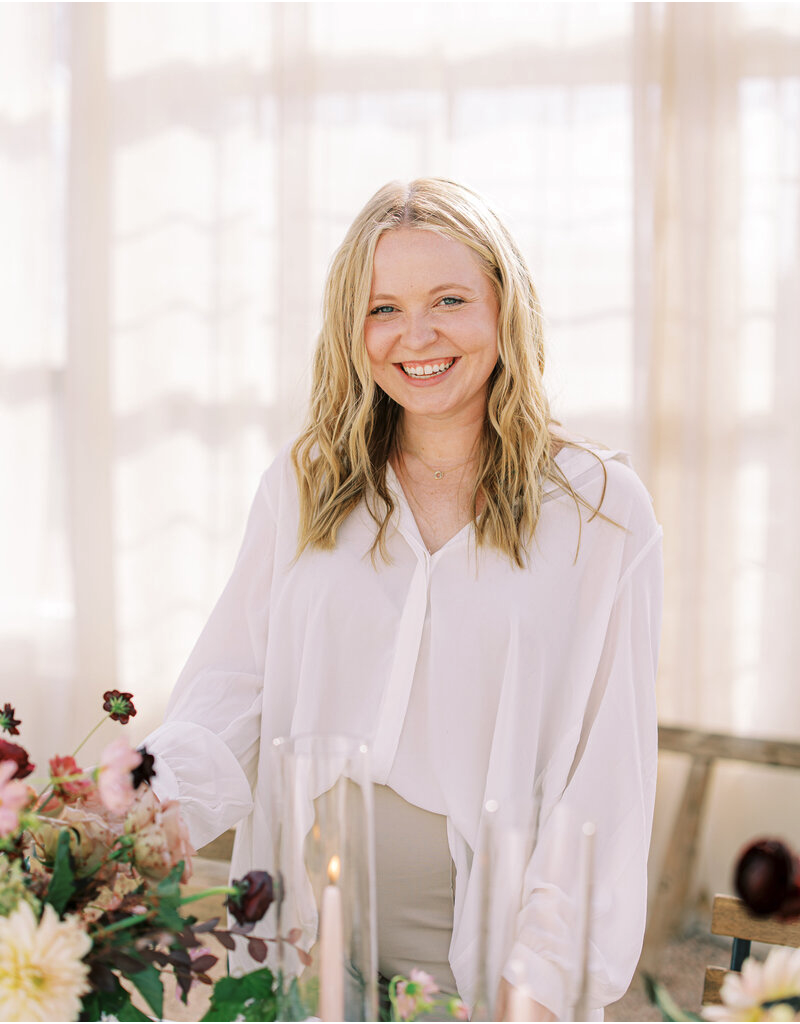 A smiling woman with blonde hair wearing a white blouse, standing indoors with a table of flowers and candles in front of her.