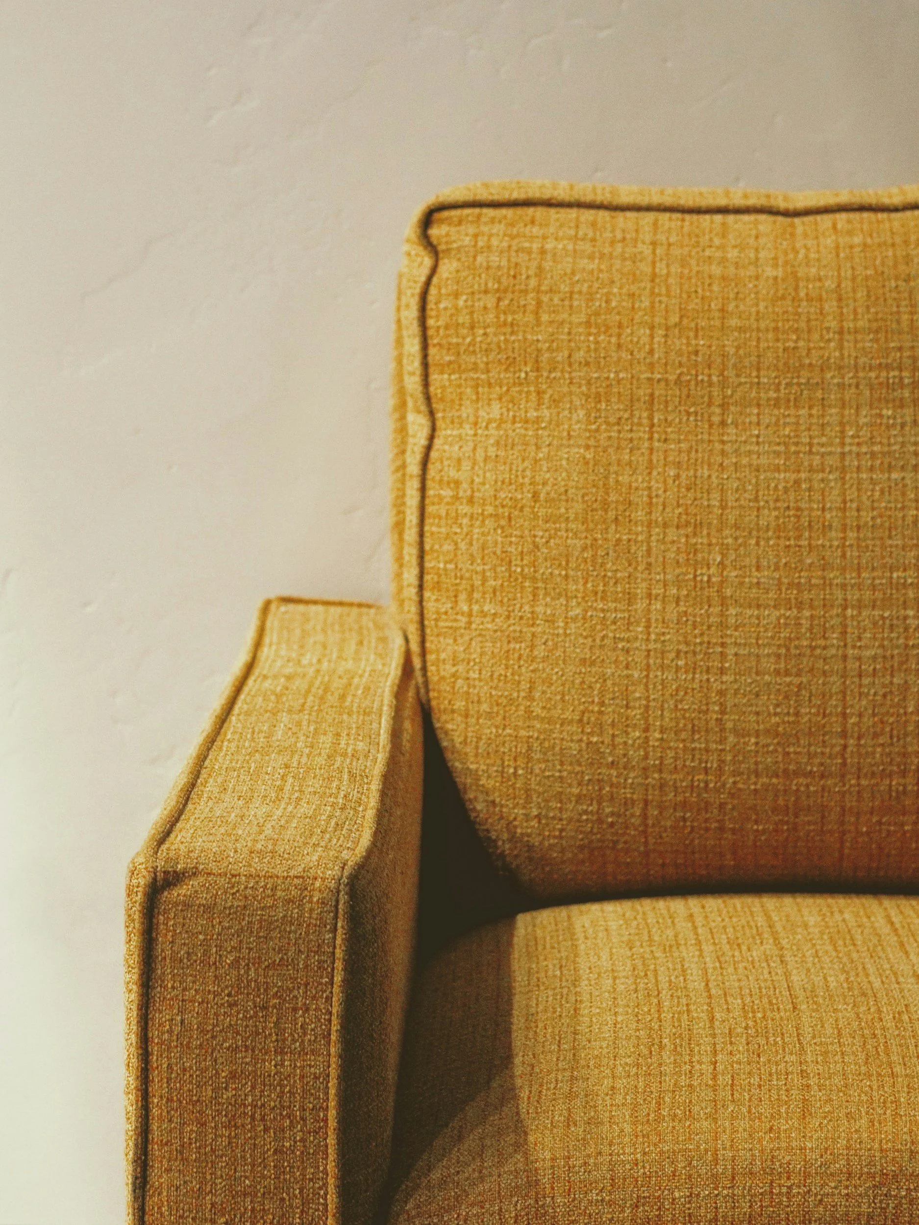 Close-up of a mustard yellow upholstered armchair with a textured fabric, black piping along the edges, against a plain white wall.