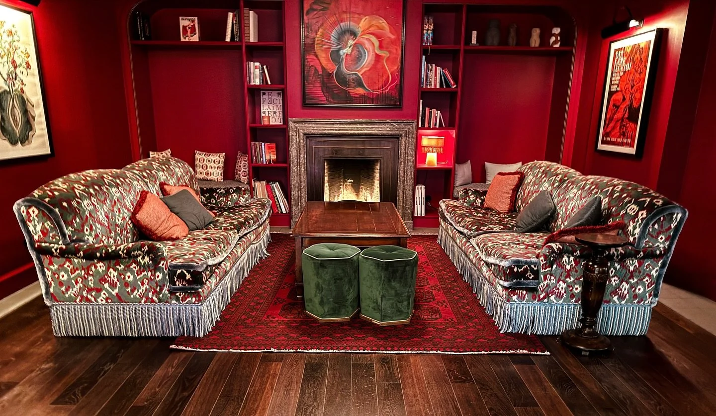 An elegant living room featuring two vintage floral-patterned sofas with throw pillows, a wooden coffee table, and a fireplace set against a deep red wall with built-in bookshelves, paintings, and a vibrant red area rug on a hardwood floor.