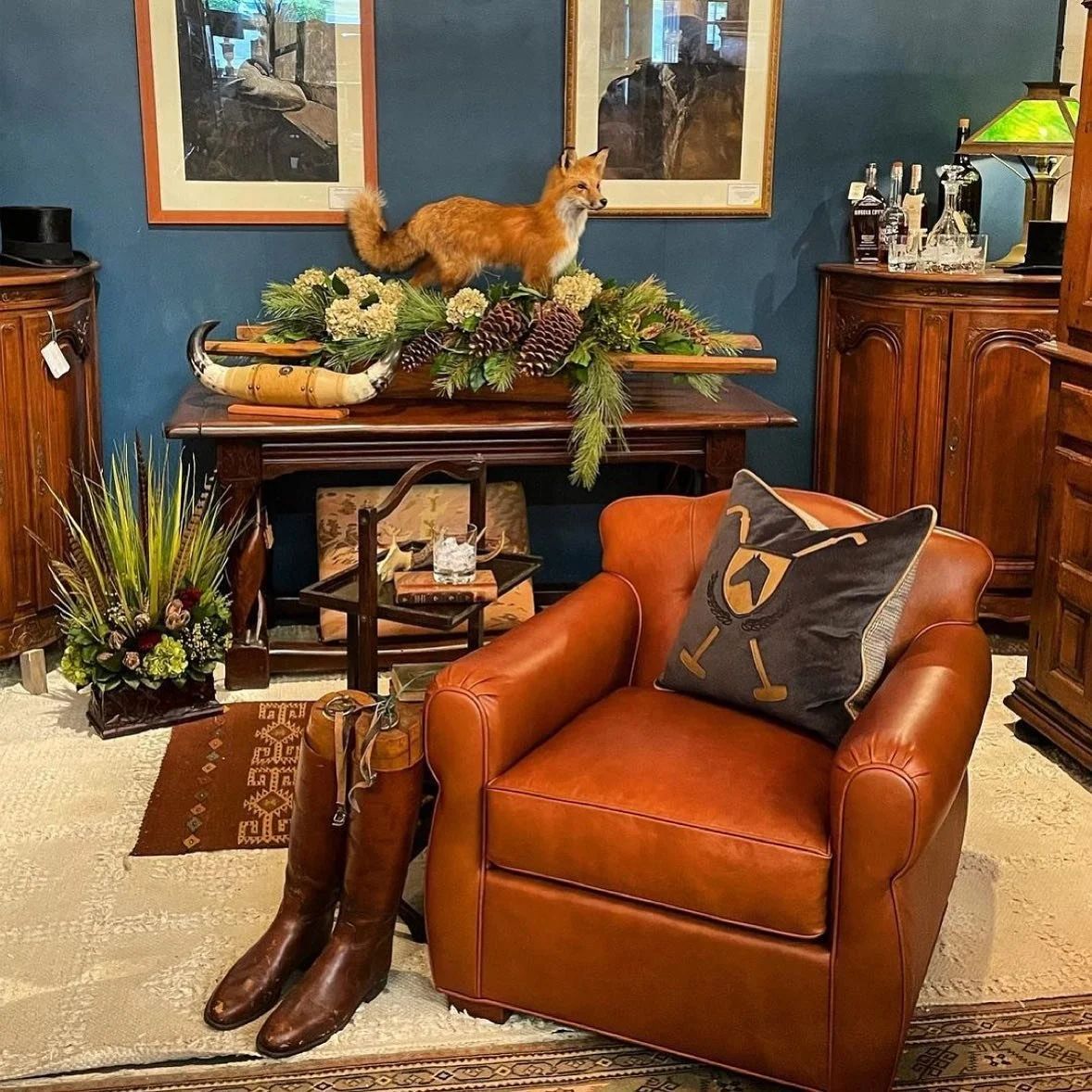 A stuffed fox statue on a wooden table decorated with pinecones and greenery in a living room. A pair of brown cowboy boots in front of a leather armchair with a pillow. Barware and artwork are on nearby furniture.