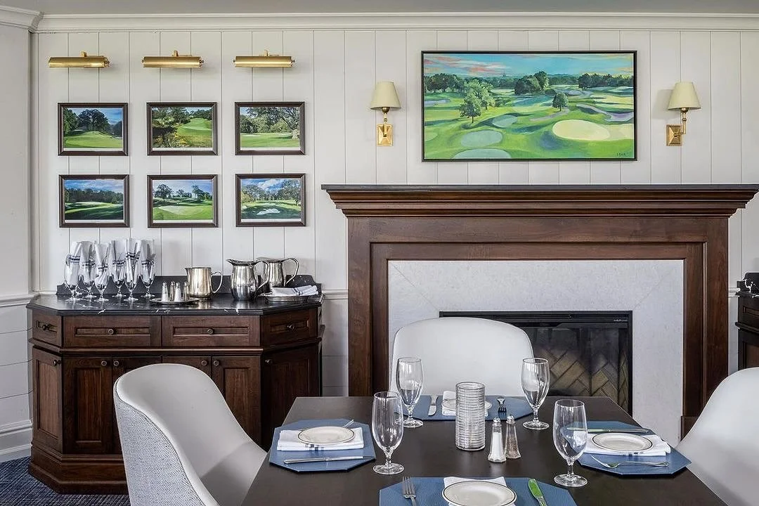 Elegant dining room with a dark wood table set with glasses, plates, and utensils, surrounded by white chairs. A fireplace with a wooden mantel and framed artwork of a golf course above it, flanked by wall-mounted lamps. A side cabinet with glassware