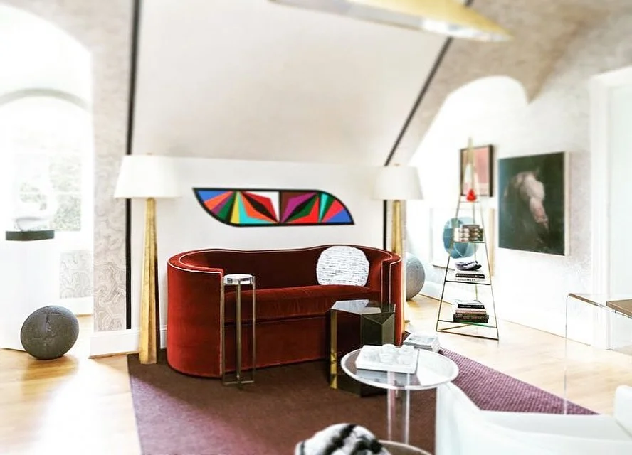 Interior of a modern living room with a red velvet sofa, colorful abstract art above it, white lamps on tall gold stands, a white marble coffee table with books, a black side table, and a white armchair. There is a large window, a brown area rug, and