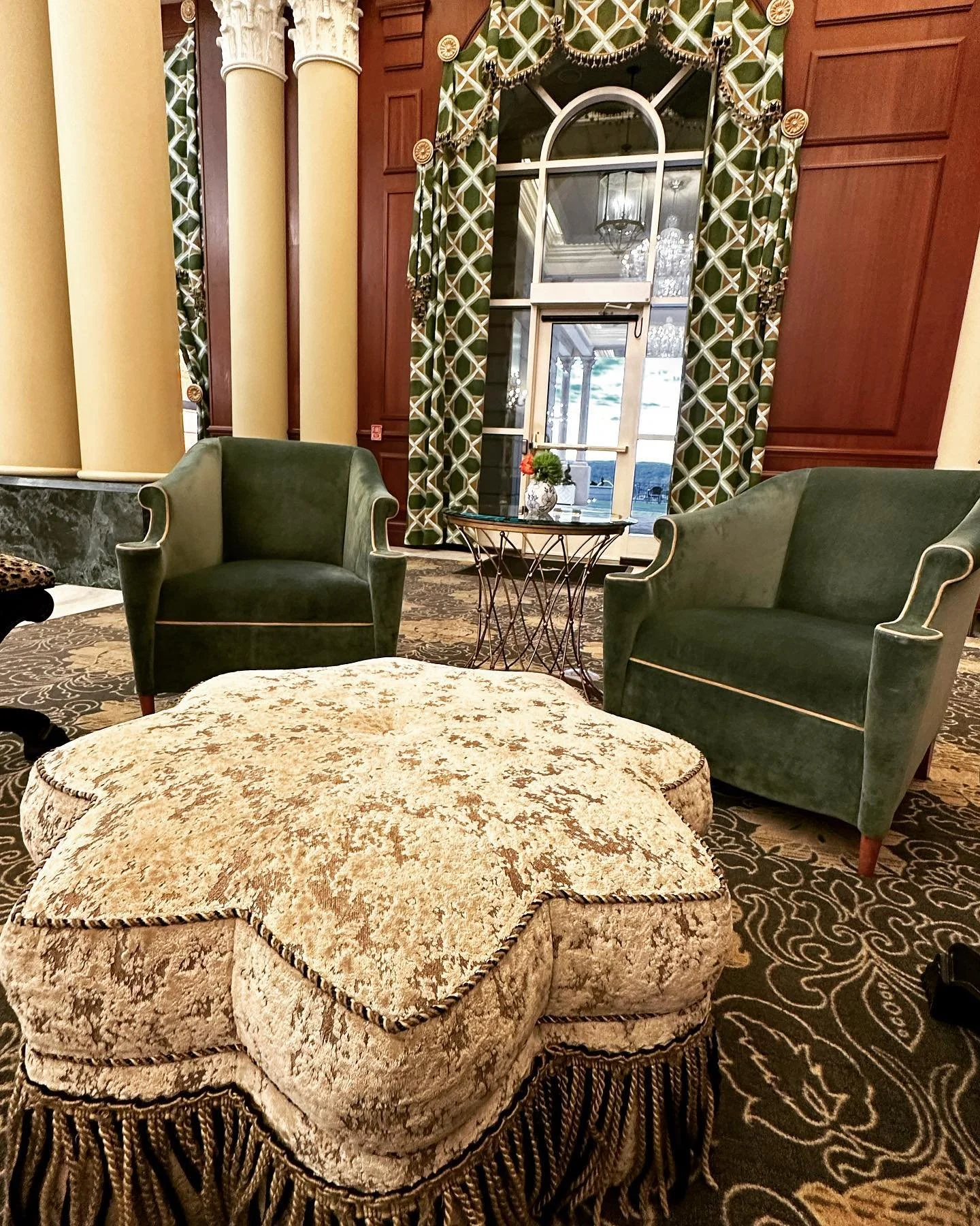 Two green upholstered armchairs near a window with ornate green and white curtains, a small round table with a decorative plant, and a patterned carpet in a luxurious room with tall columns.