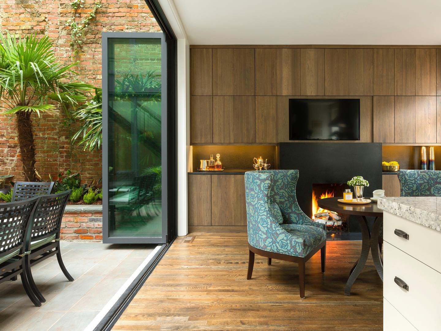 Interior of a modern living space with a fireplace, a flat-screen TV, a round table with a patterned chair, and an outdoor patio with a brick wall, green plants, and black chairs.