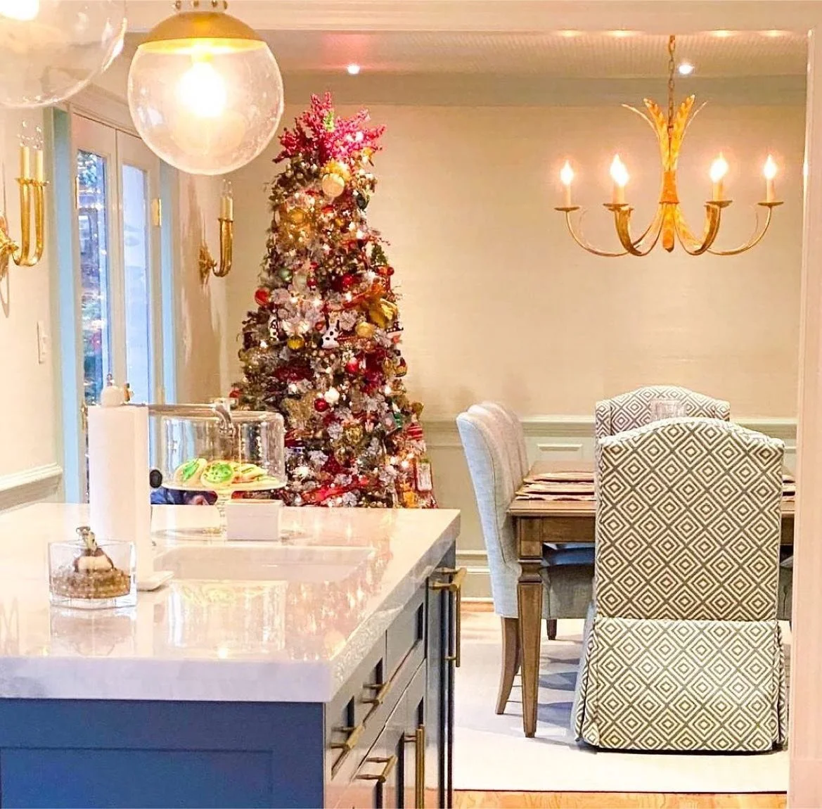 A decorated Christmas tree in a dining room with a chandelier and wall-mounted lights, next to a table with chairs, with a view outside through windows.