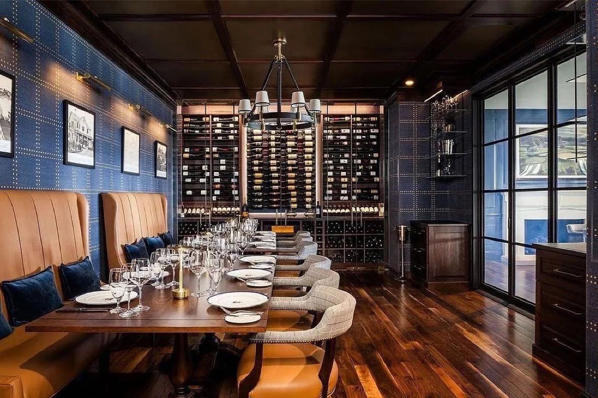 Elegant dining room with a wooden table set with glasses, plates, and napkins, surrounded by upholstered chairs. Wall-mounted artwork and a wine rack are visible, with dark wood floors and a large window.