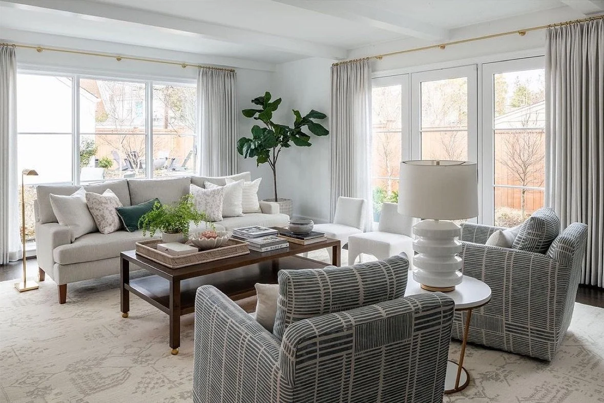 Bright living room with large windows, white curtains, white sofa with multiple pillows, gray patterned armchairs, a wooden coffee table with books and decorative items, a lamp with a white shade, and a potted plant in the corner.