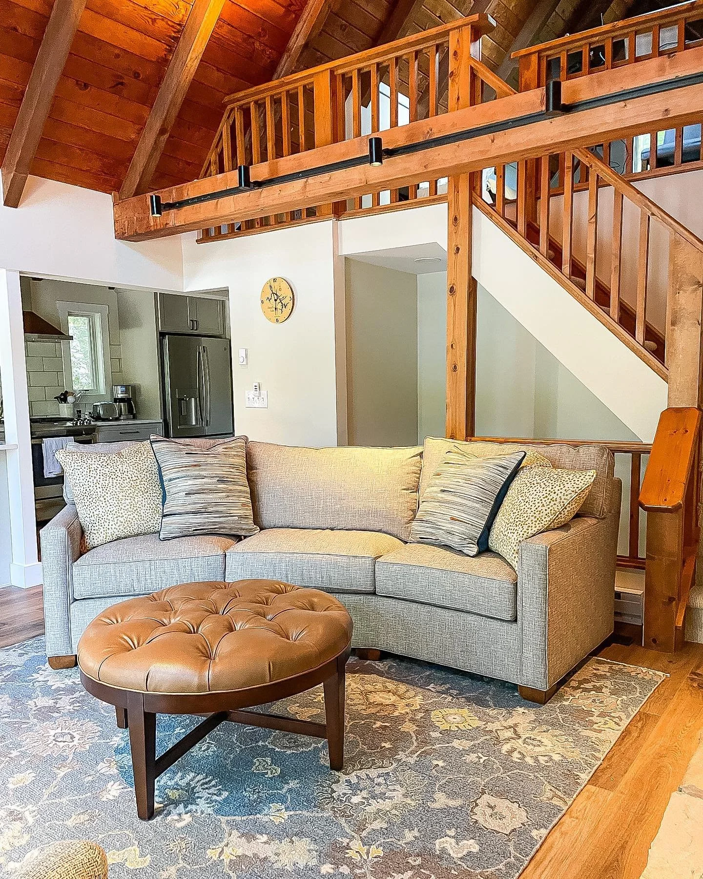 Living room with beige sofa and patterned pillows, tufted brown leather ottoman, area rug, wooden staircase and balcony with wood railing, open to kitchen with stainless steel refrigerator, coffee maker, and white tiled backsplash.
