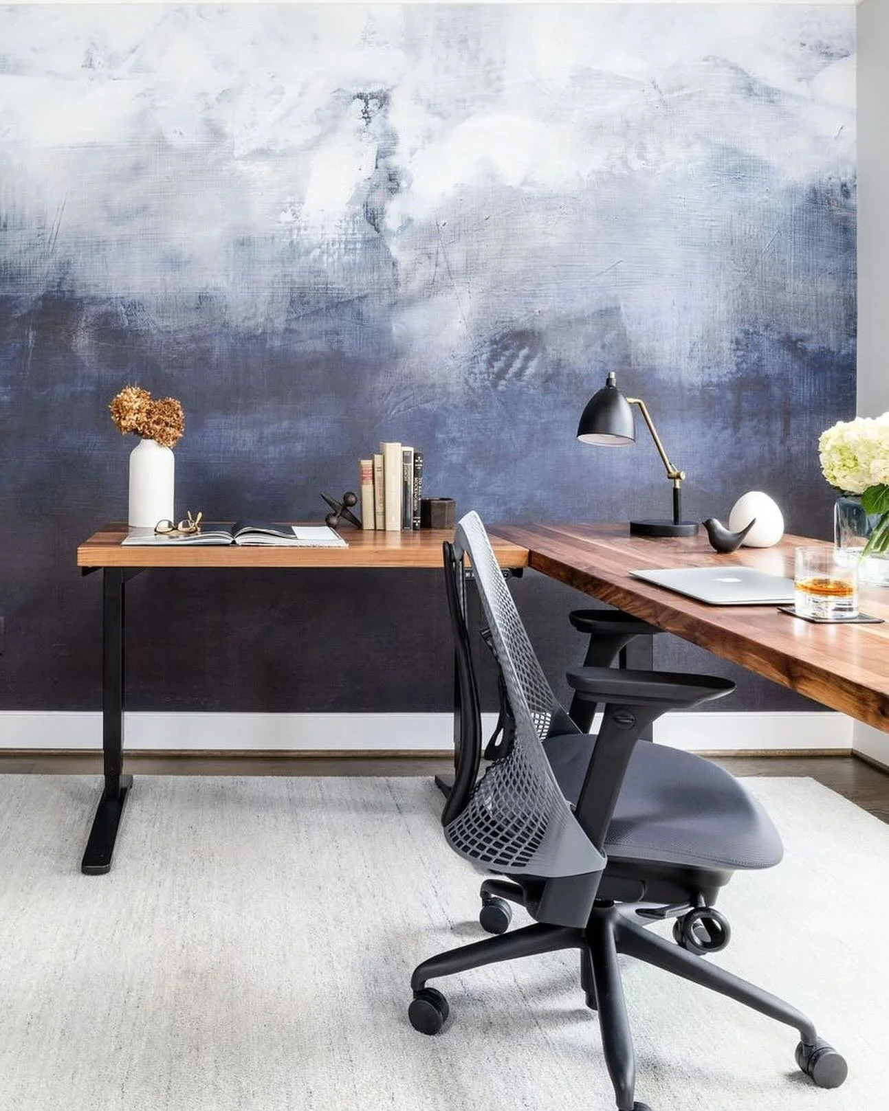 Modern home office with a wooden desk, black office chair, decorative objects, books, lamp, and flowers against a textured blue-gray wall.