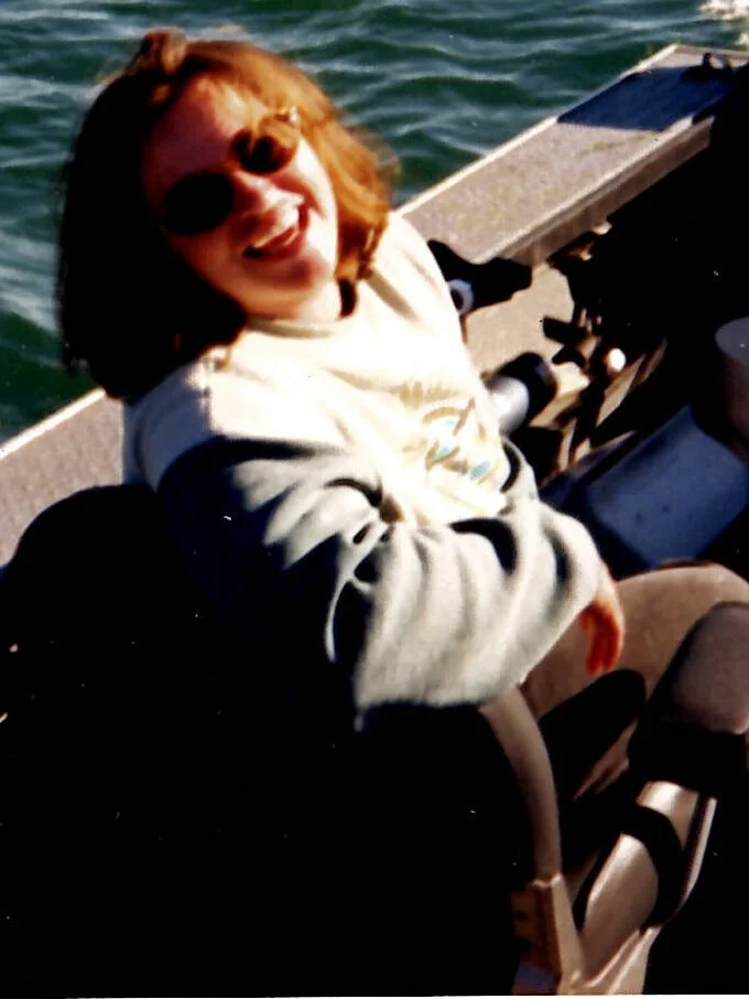 A woman with red hair and sunglasses sitting on a boat, smiling at the camera.