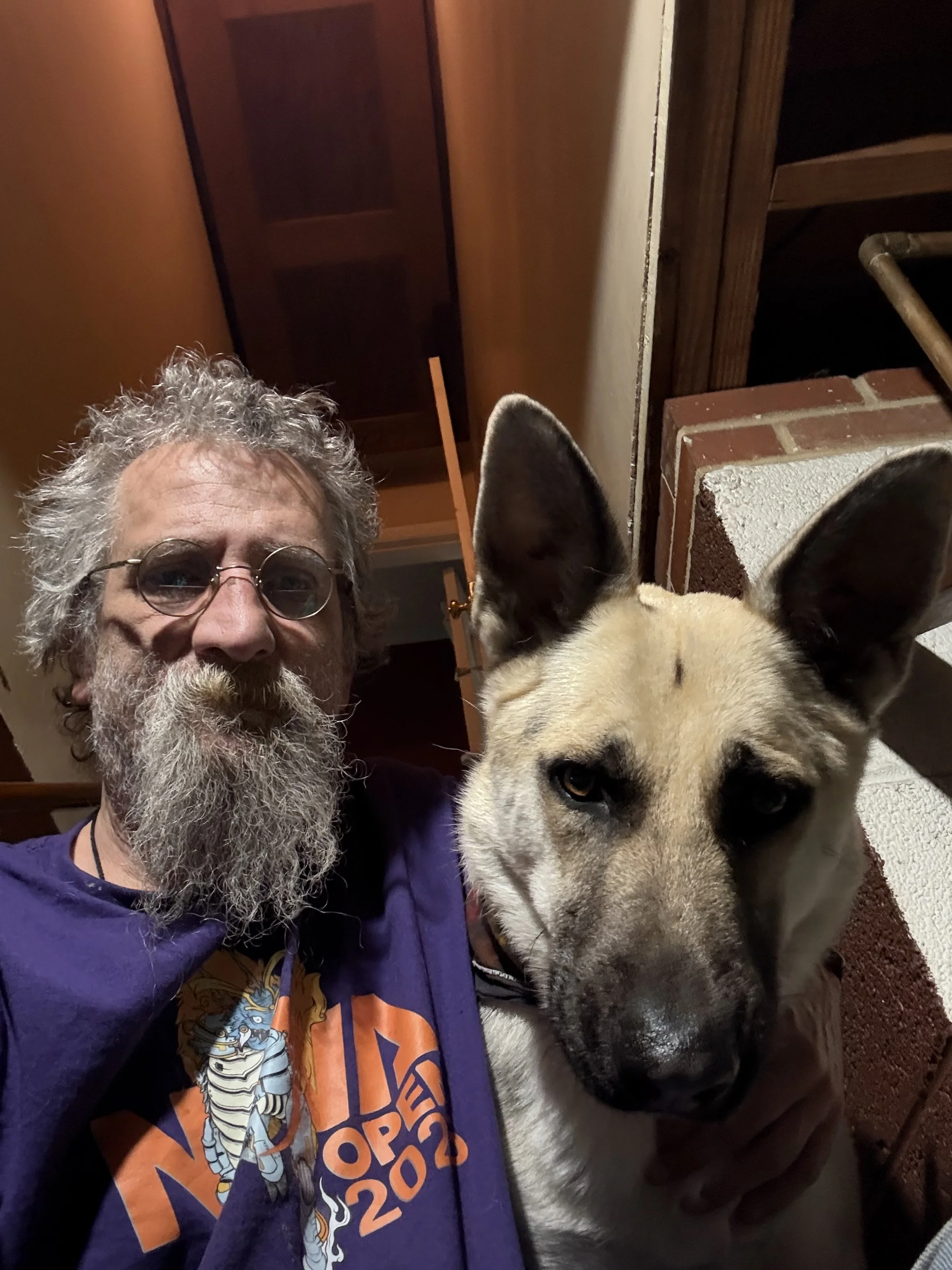 A man with a curly gray beard and glasses taking a selfie with a large dog that has a tan and black coat, pointed ears, and a serious expression inside a home with wooden and brick interior.