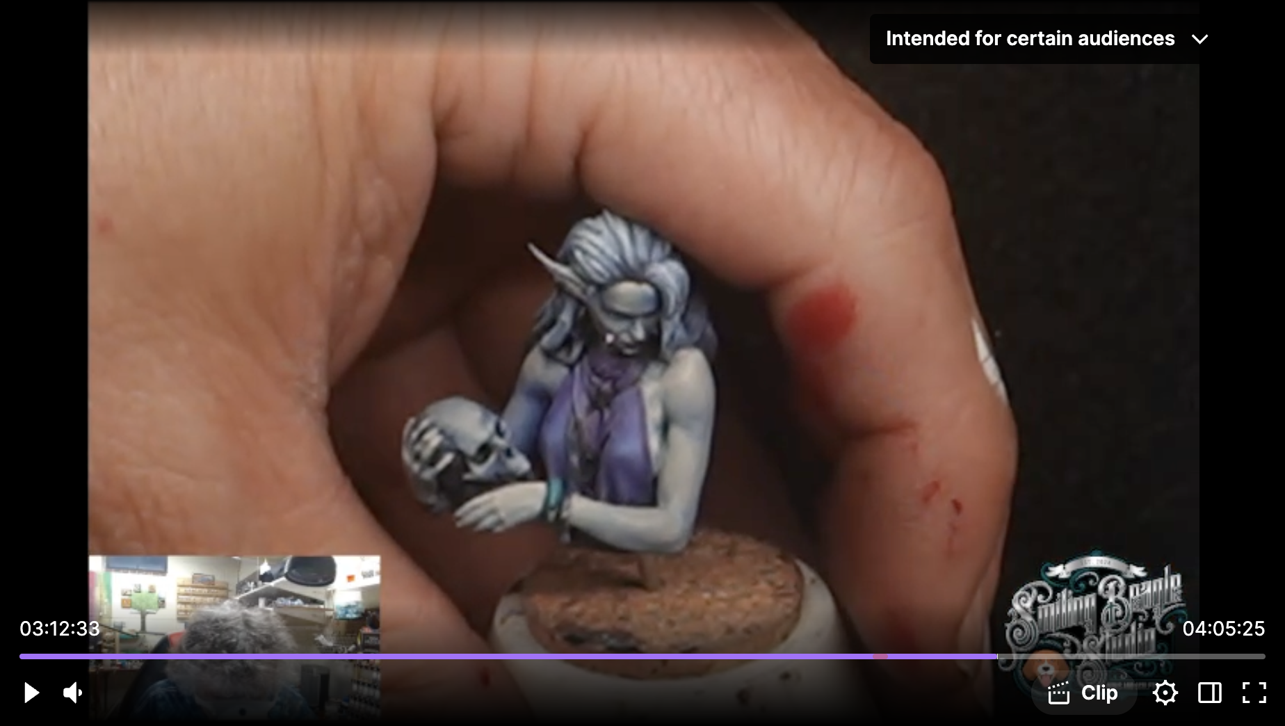A close-up of a painted miniature figure of a fantasy character with gray hair, pointed ears, and blue skin, held between a person's fingers with some bloodstains on the fingers. The miniature appears to be part of a tabletop game or collection.