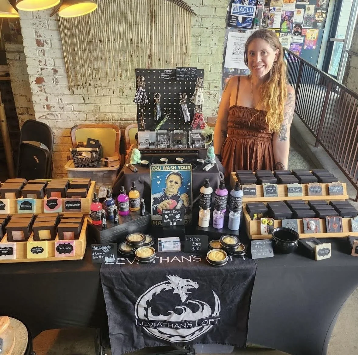 A woman standing behind a table with soap and skincare products at an indoor market. The table has a black cloth with a Leviathan's Loft banner, and various soaps, lotions, and oils are displayed. Behind her is a pegboard and there are chairs and a brick wall in the background.
