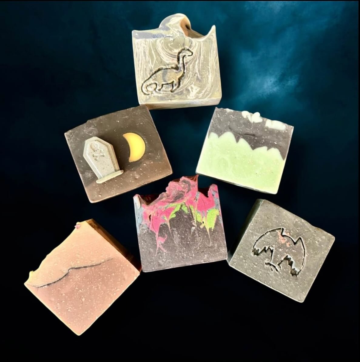An assortment of six colorful, patterned soap bars with various designs, including an elephant, a moon with a tent, mountains, a pink and green splash, a skull, and a raised surface with a lighter pink hue, arranged on a dark blue surface.