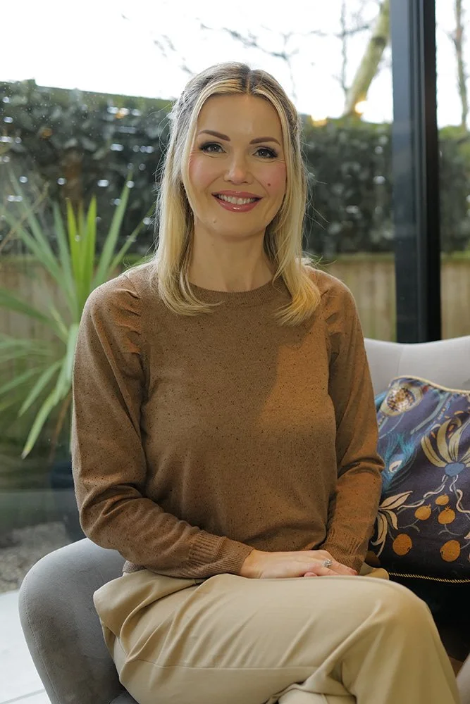 A smiling woman with blonde hair sitting on a gray chair indoors, with a large window and garden visible in the background.