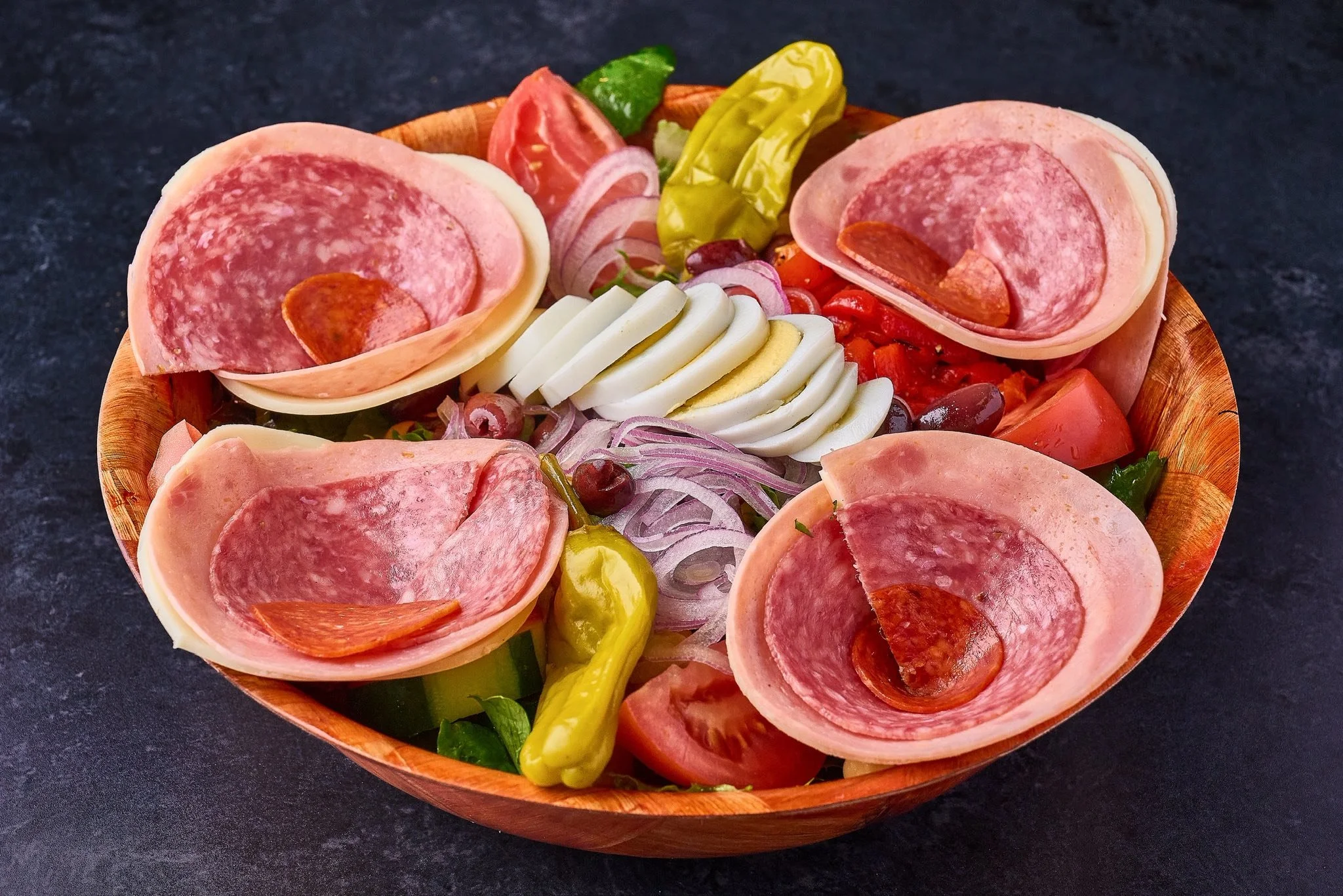 A wooden bowl filled with slices of salami, cherry tomatoes, sliced hard-boiled eggs, red onion, green peppers, and black and green olives, arranged as a salad on a dark surface.