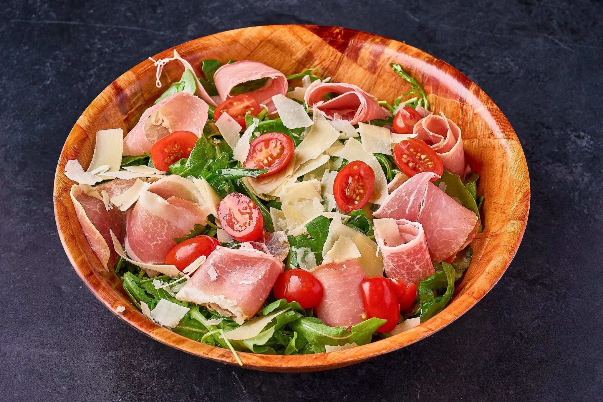 Fresh salad with cherry tomatoes, slices of cheese, prosciutto, and arugula in a wooden bowl on a dark surface.