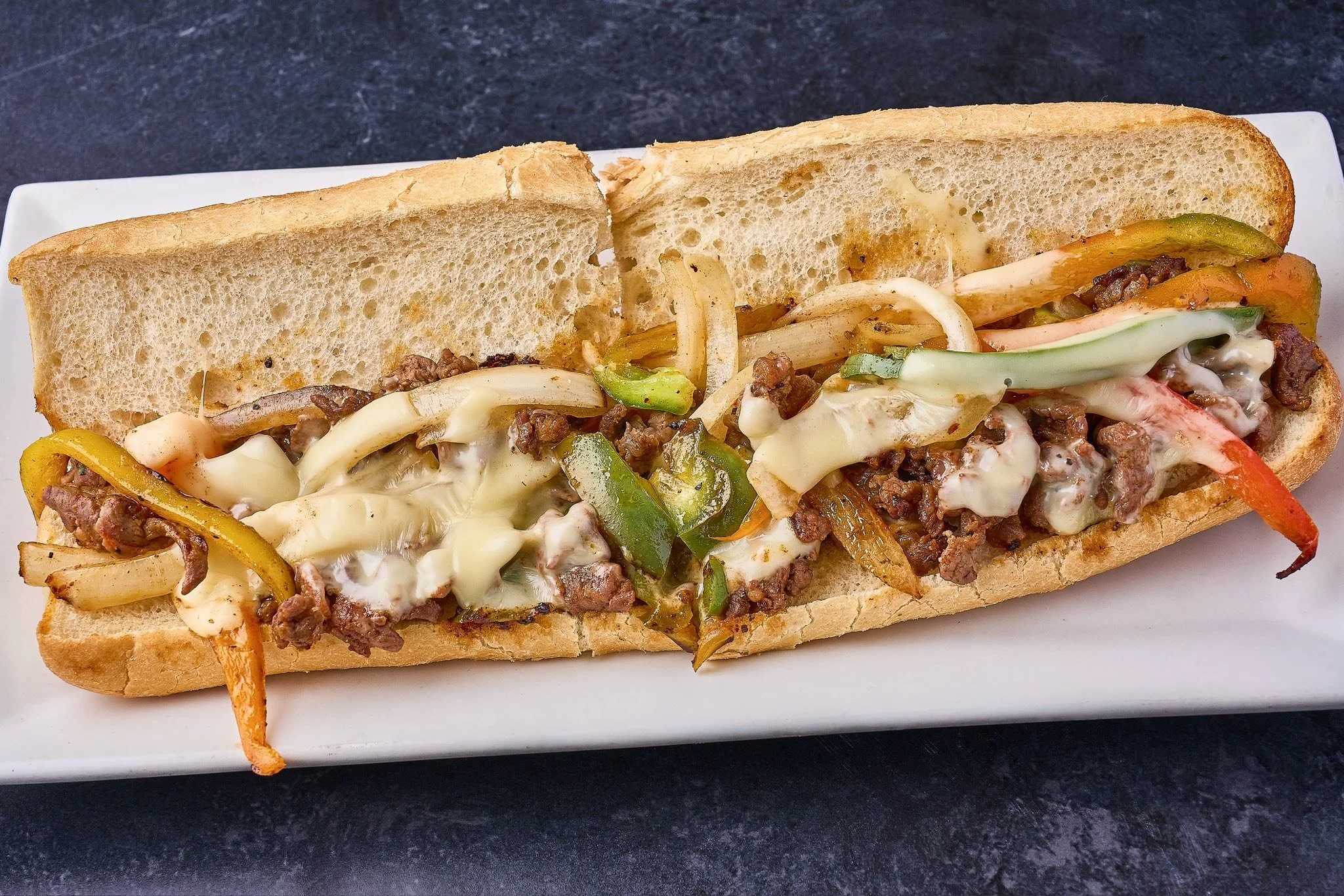A sub sandwich with sliced bread filled with ground beef, melted cheese, and sautéed vegetables including onions, bell peppers, and possibly zucchini or pickles, served on a white rectangular plate.