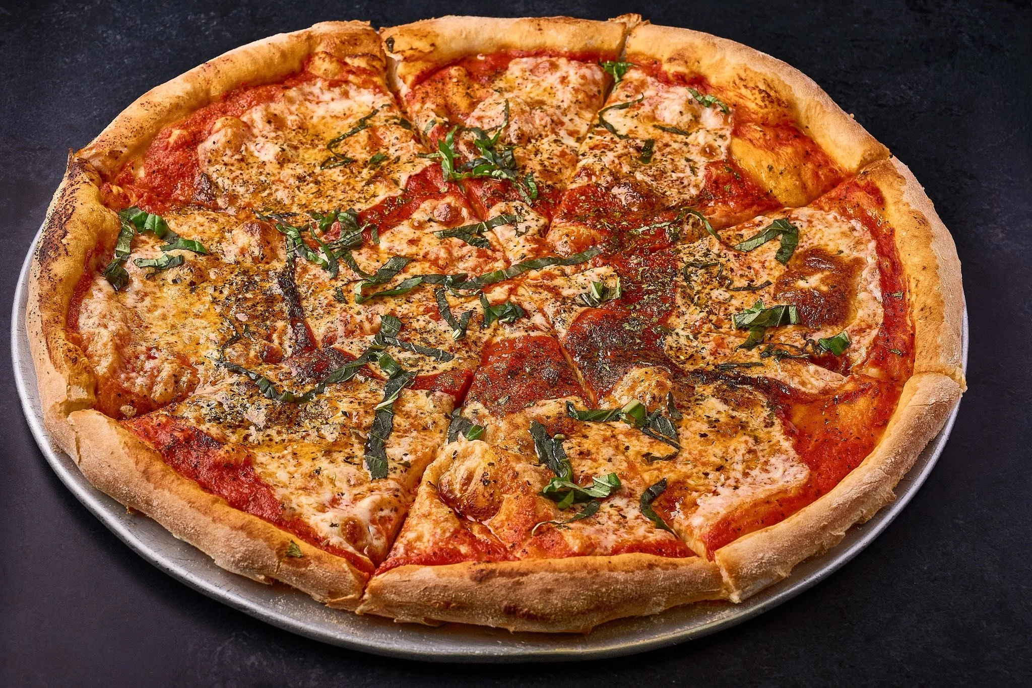 A pepperoni, cheese, and basil pizza on a silver tray against a black background.