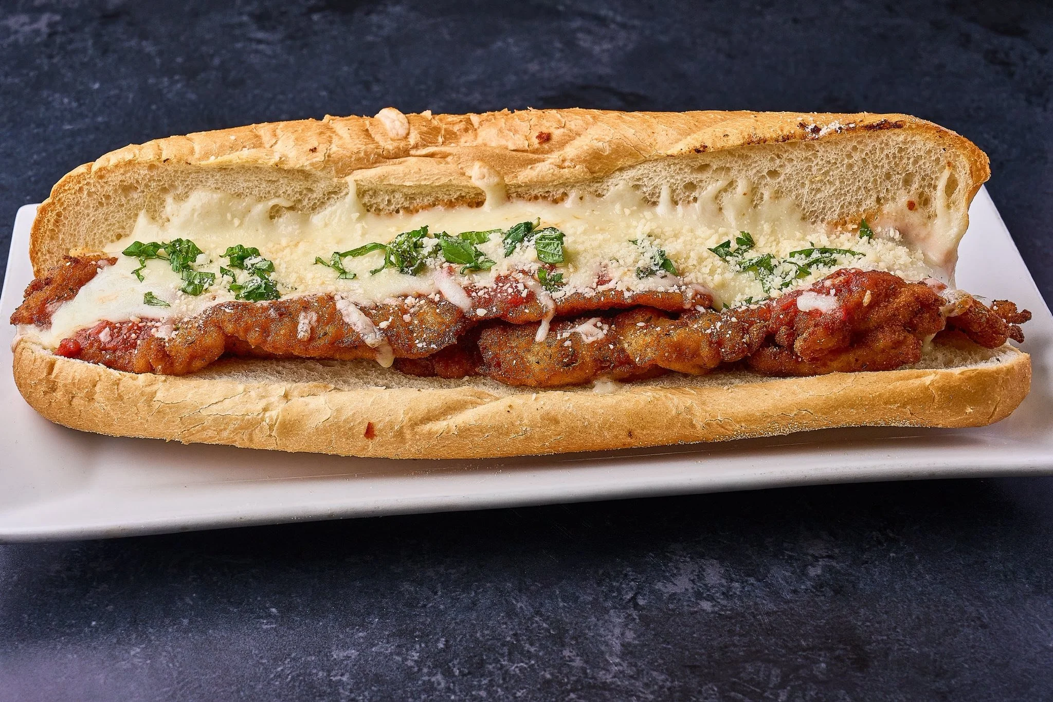 Chicken Parmesan sub sandwich with fried chicken, melted cheese, marinara sauce, and chopped herbs in a hoagie roll on a white plate.