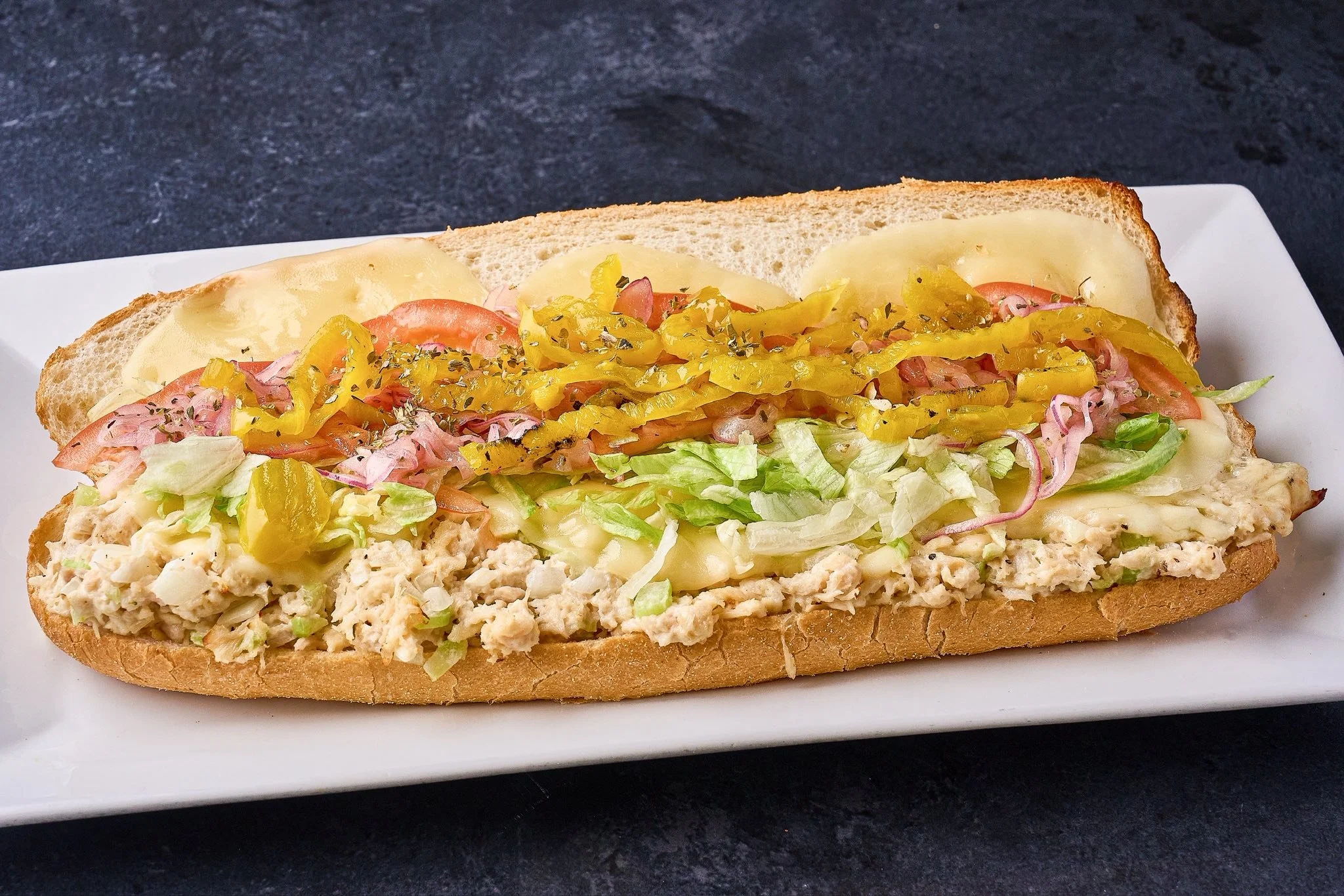 A submarine sandwich on a white plate with lettuce, tomato, pickles, shredded cabbage, shredded chicken salad, melted cheese, and yellow peppers on a hoagie bun.