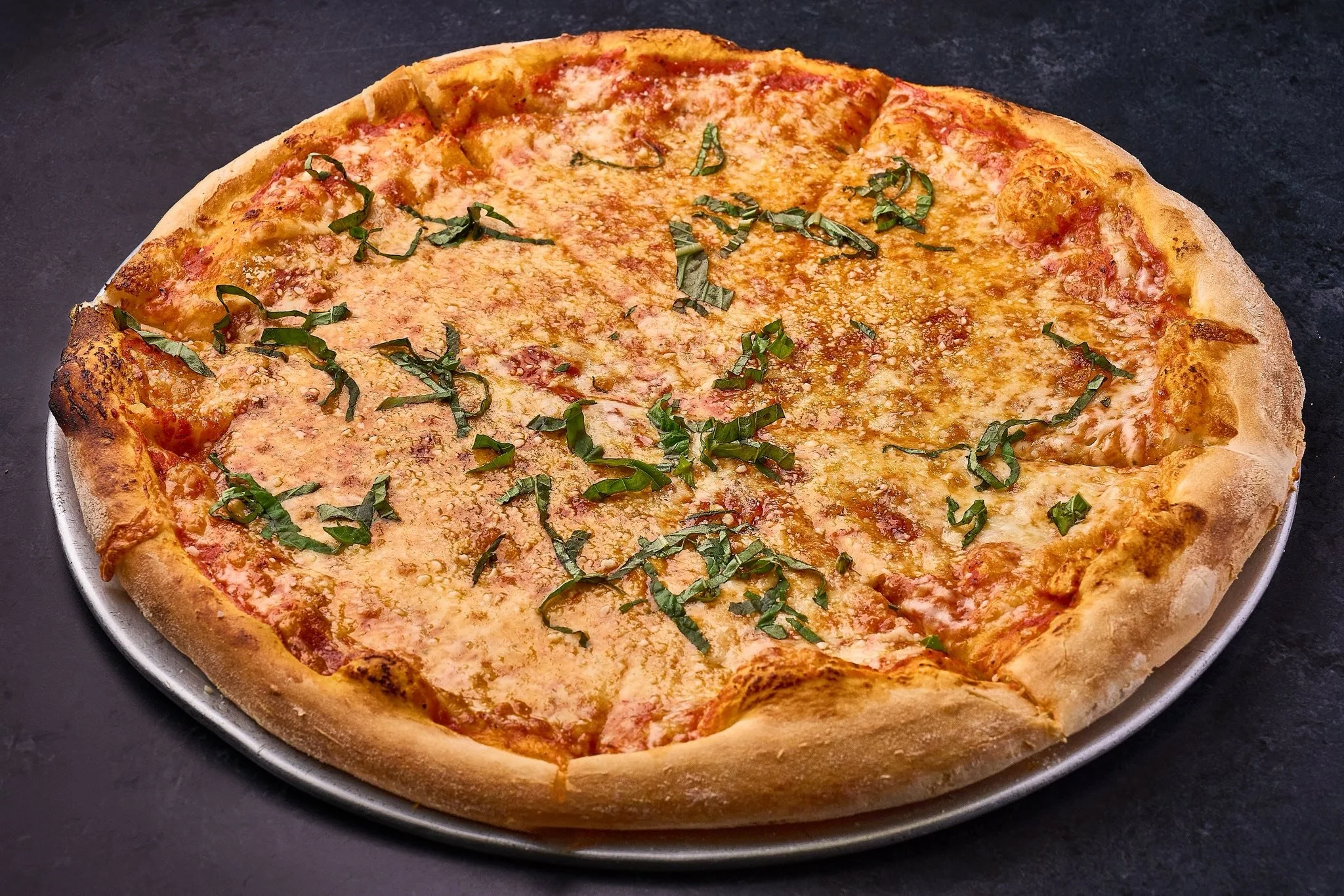 Cheese pizza with basil on a metal tray on a black surface.