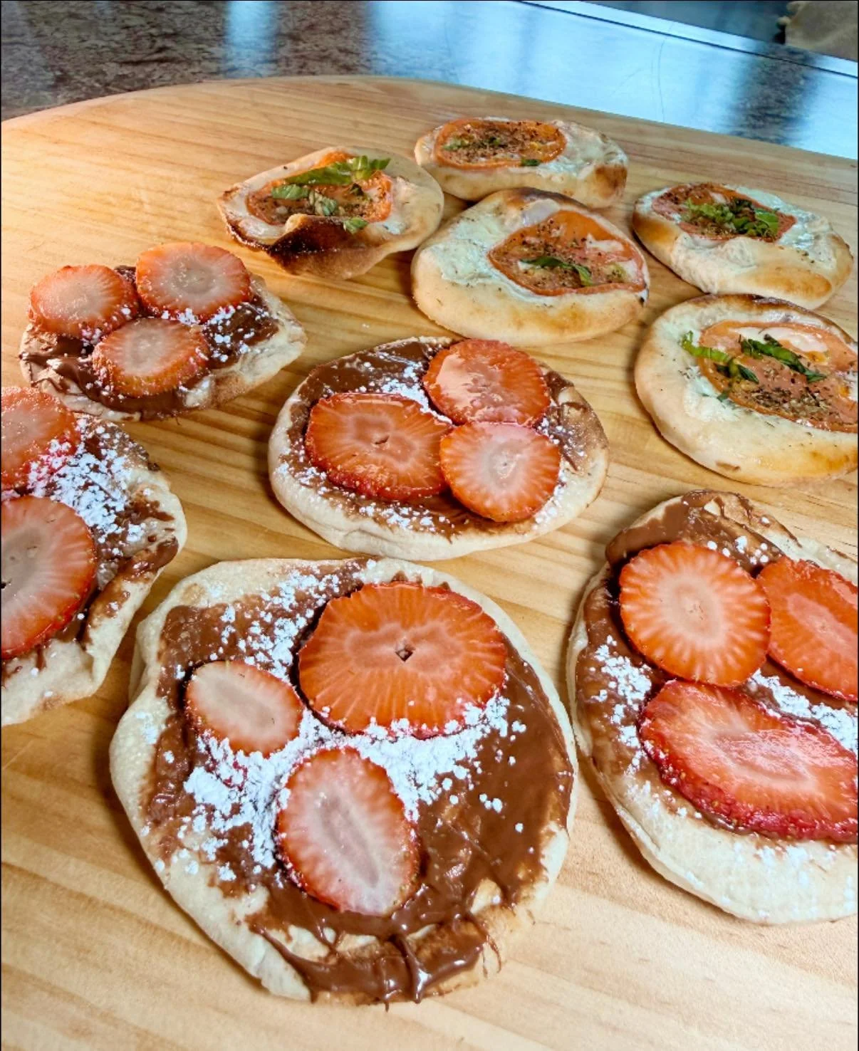 Assorted mini flatbread pizzas topped with strawberries, chocolate spread, and powdered sugar, served on a wooden cutting board.