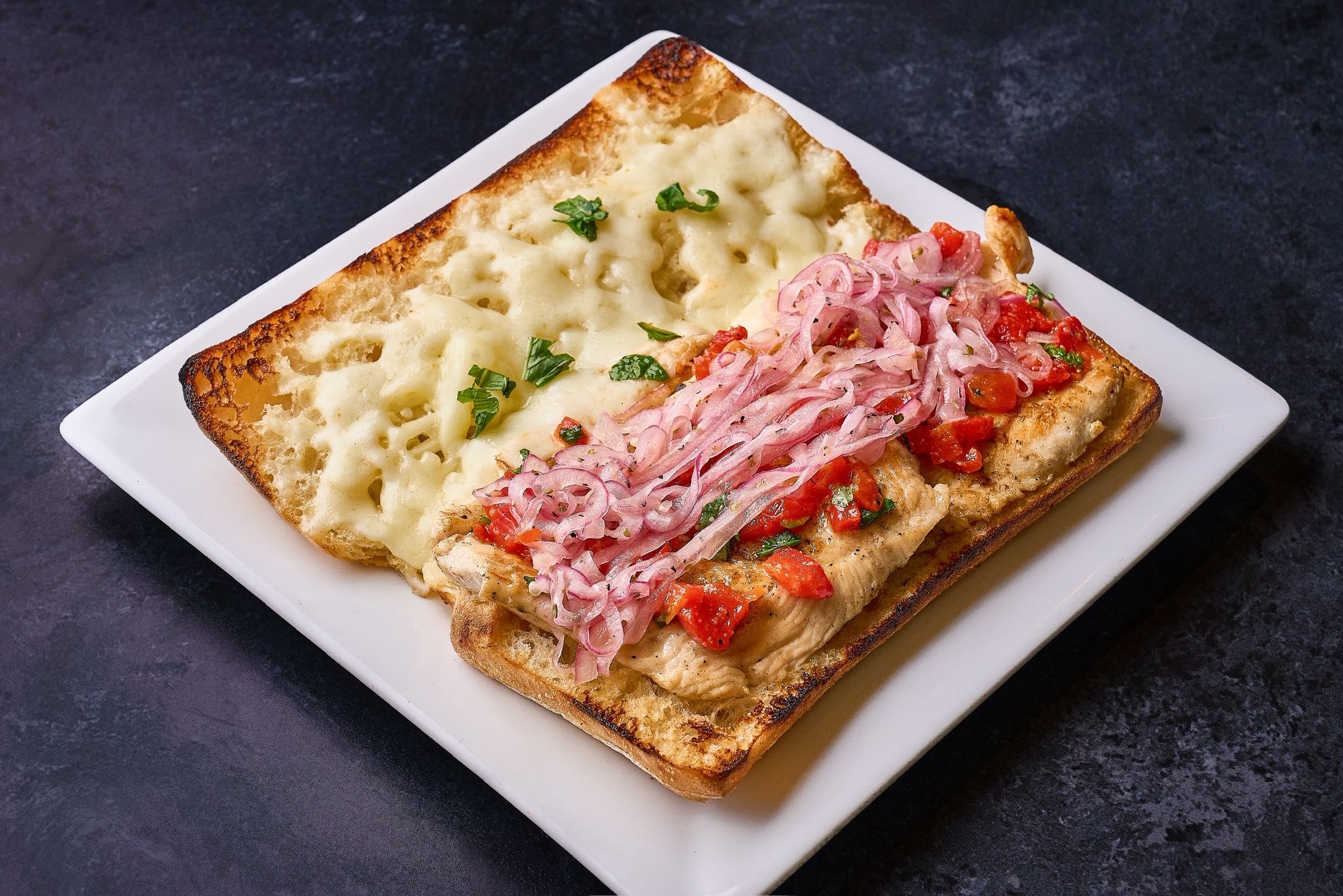 Toasted bread topped with melted cheese and chopped fresh herbs on one side, and grilled fish with tomato, onion, and herbs on the other side, served on a white plate.