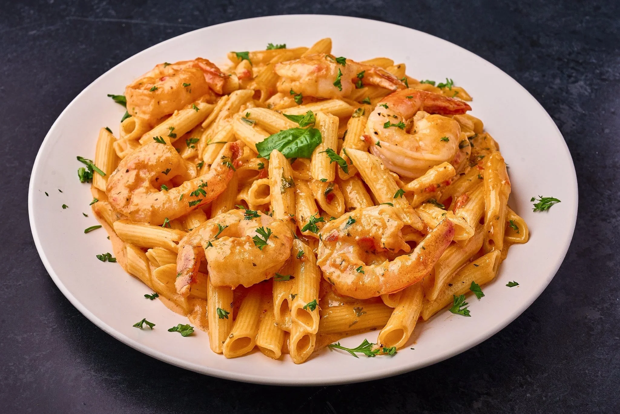 A white plate of pasta with shrimp and creamy sauce, garnished with chopped parsley on a dark surface.