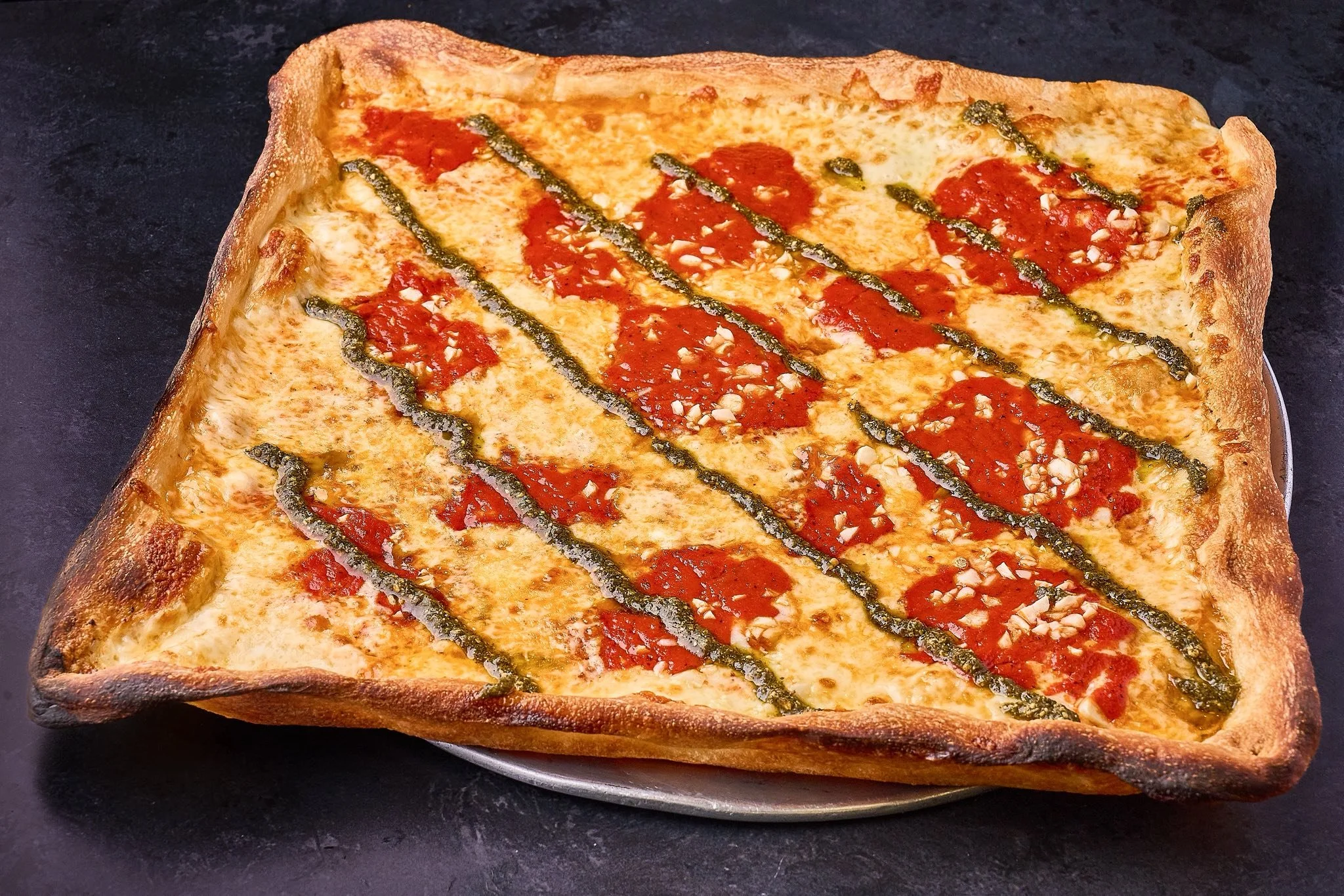 Square pizza with cheese, tomato sauce, green pesto lines, and chopped garlic on a dark surface