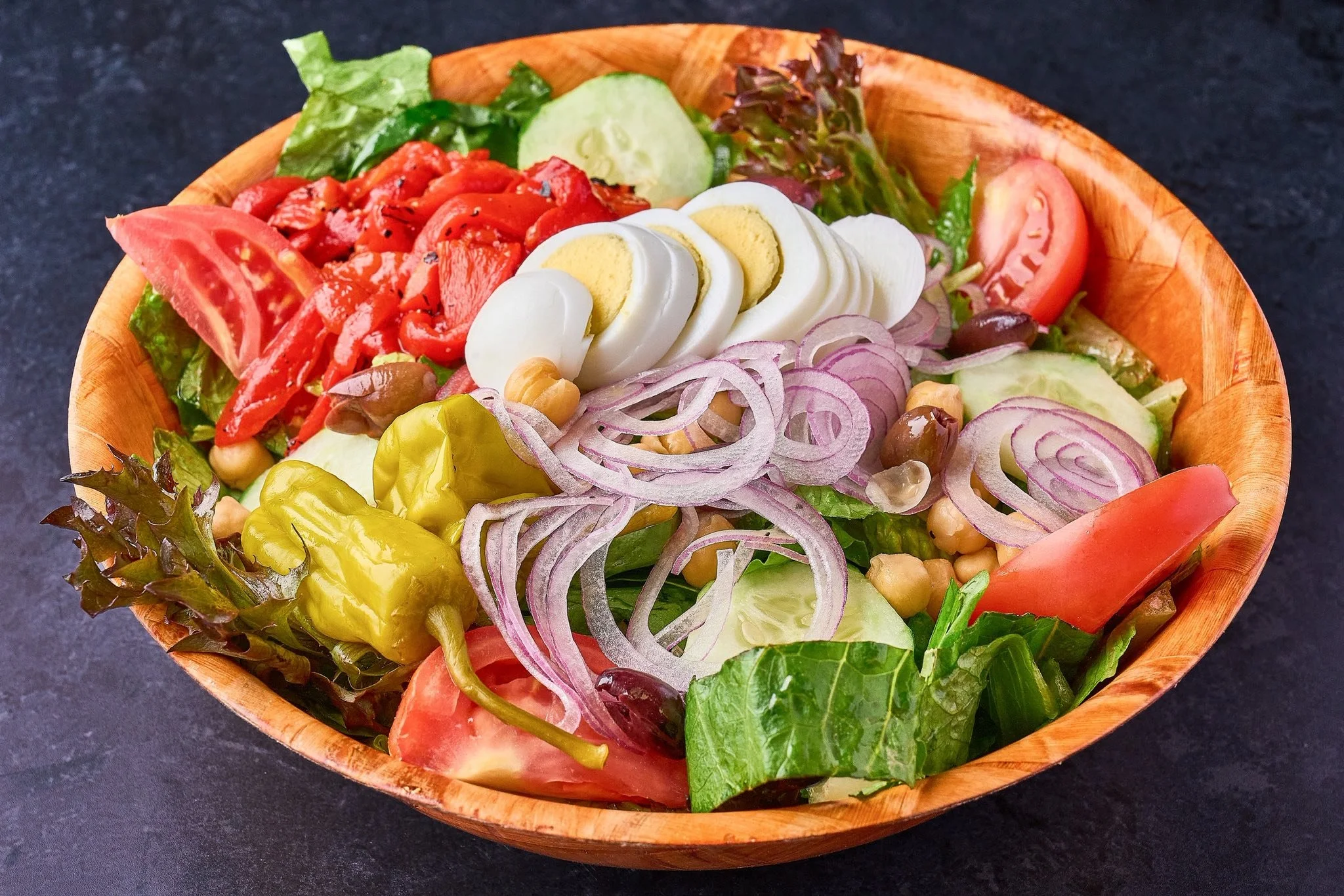 Mixed salad in a wooden bowl with lettuce, sliced tomatoes, cucumbers, onion slices, sliced boiled eggs, chickpeas, and peppers on a dark surface.