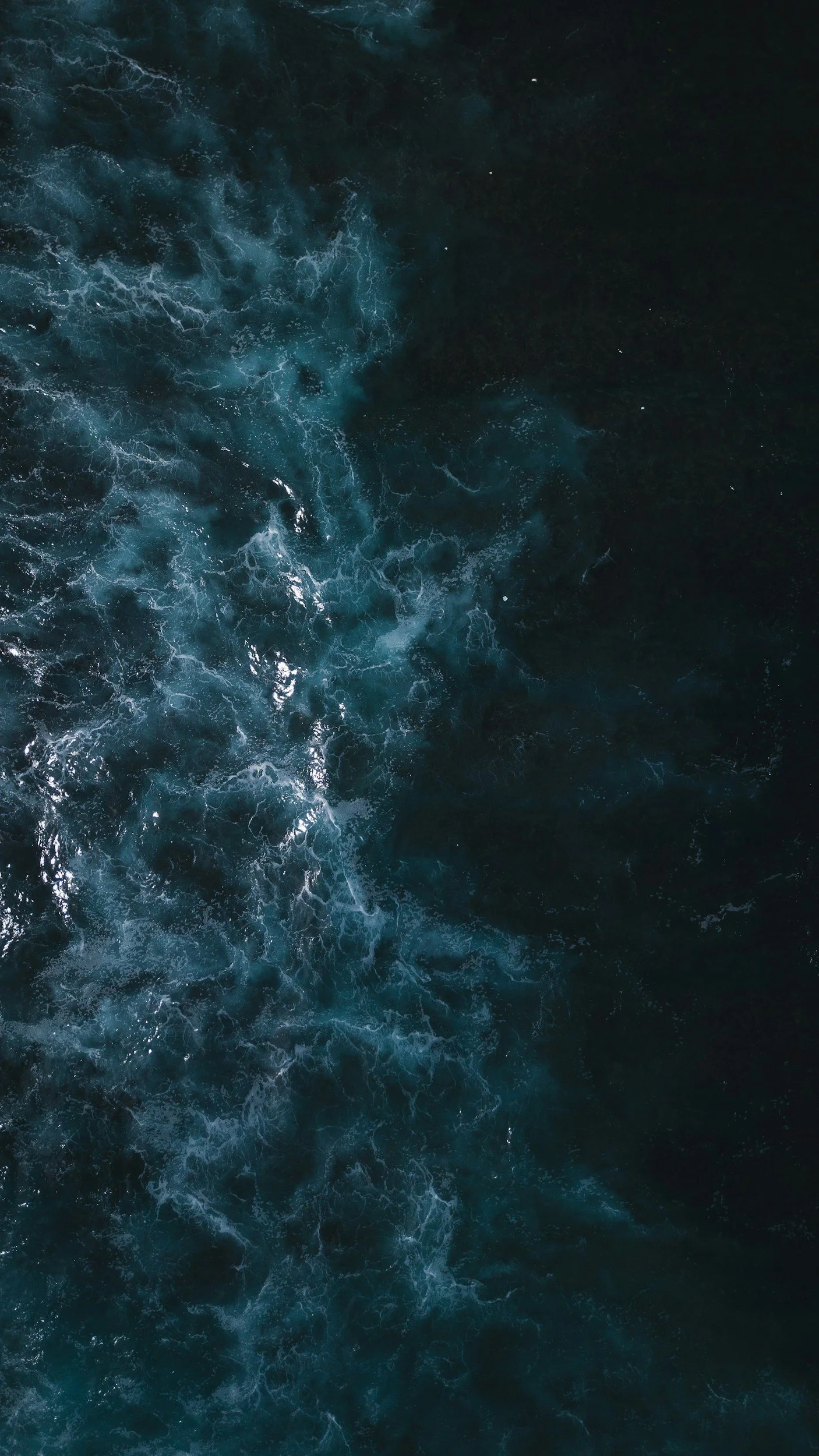 An aerial view of turbulent dark blue ocean water with white foamy waves and streaks of lighter blue.