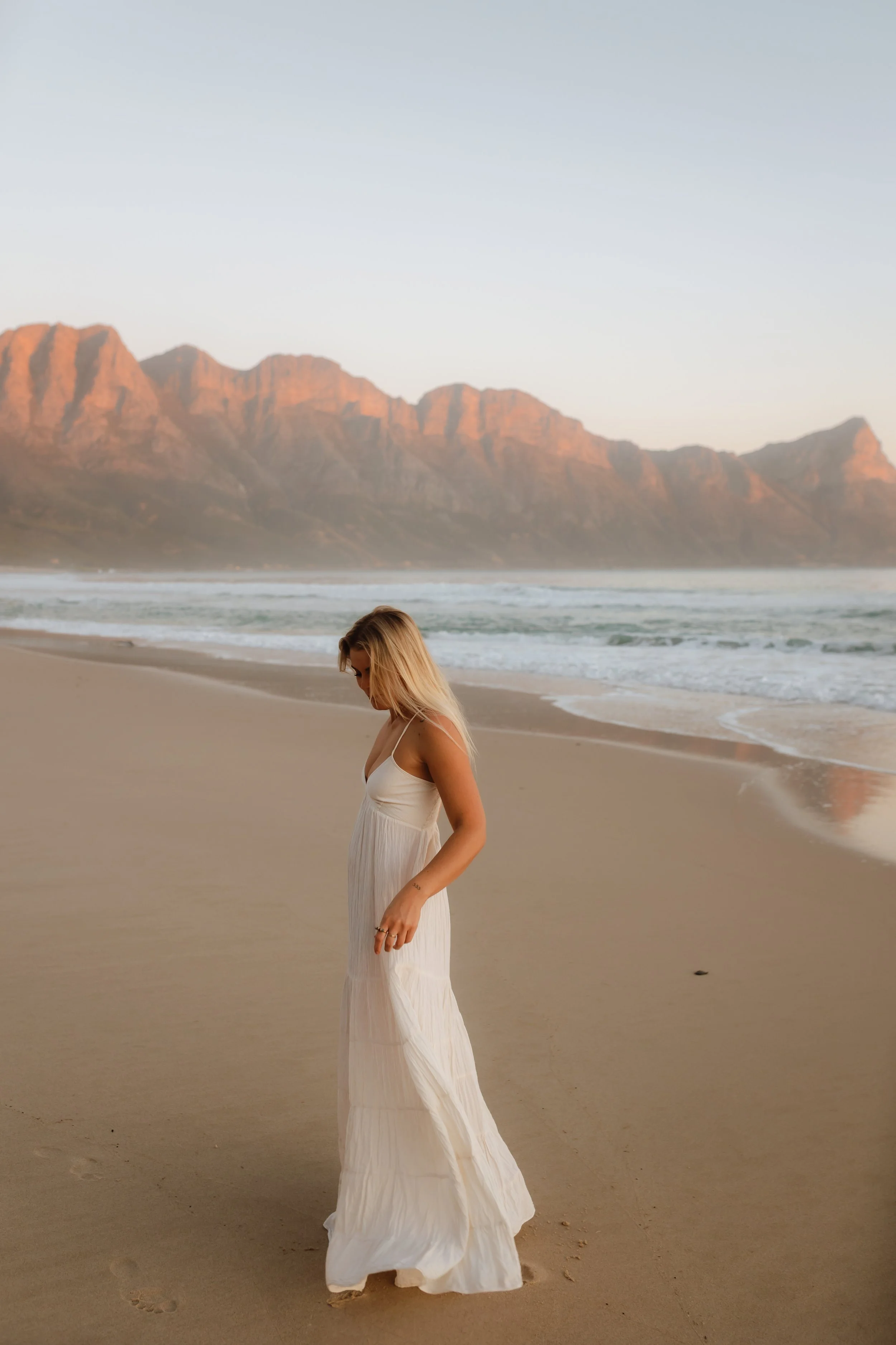 A woman in a white dress standing on a sandy beach with mountains in the background during sunset.